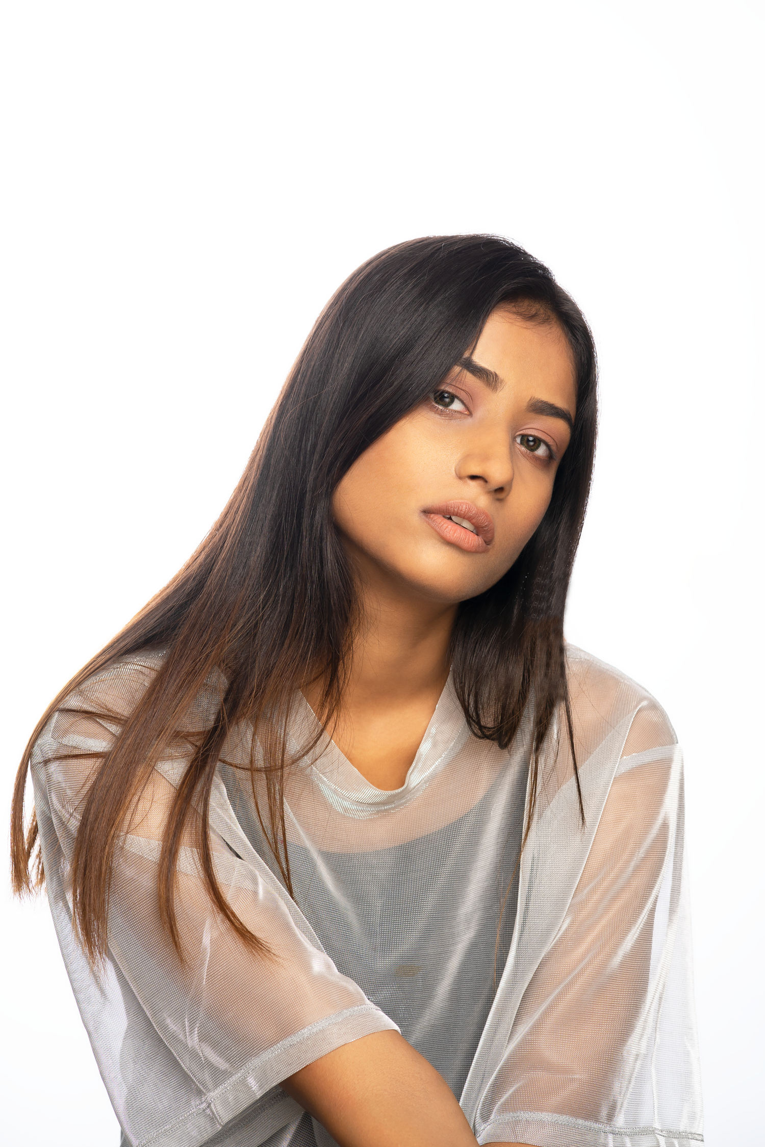Young woman with long dark hair in sheer gray top gazes softly at camera against white background.