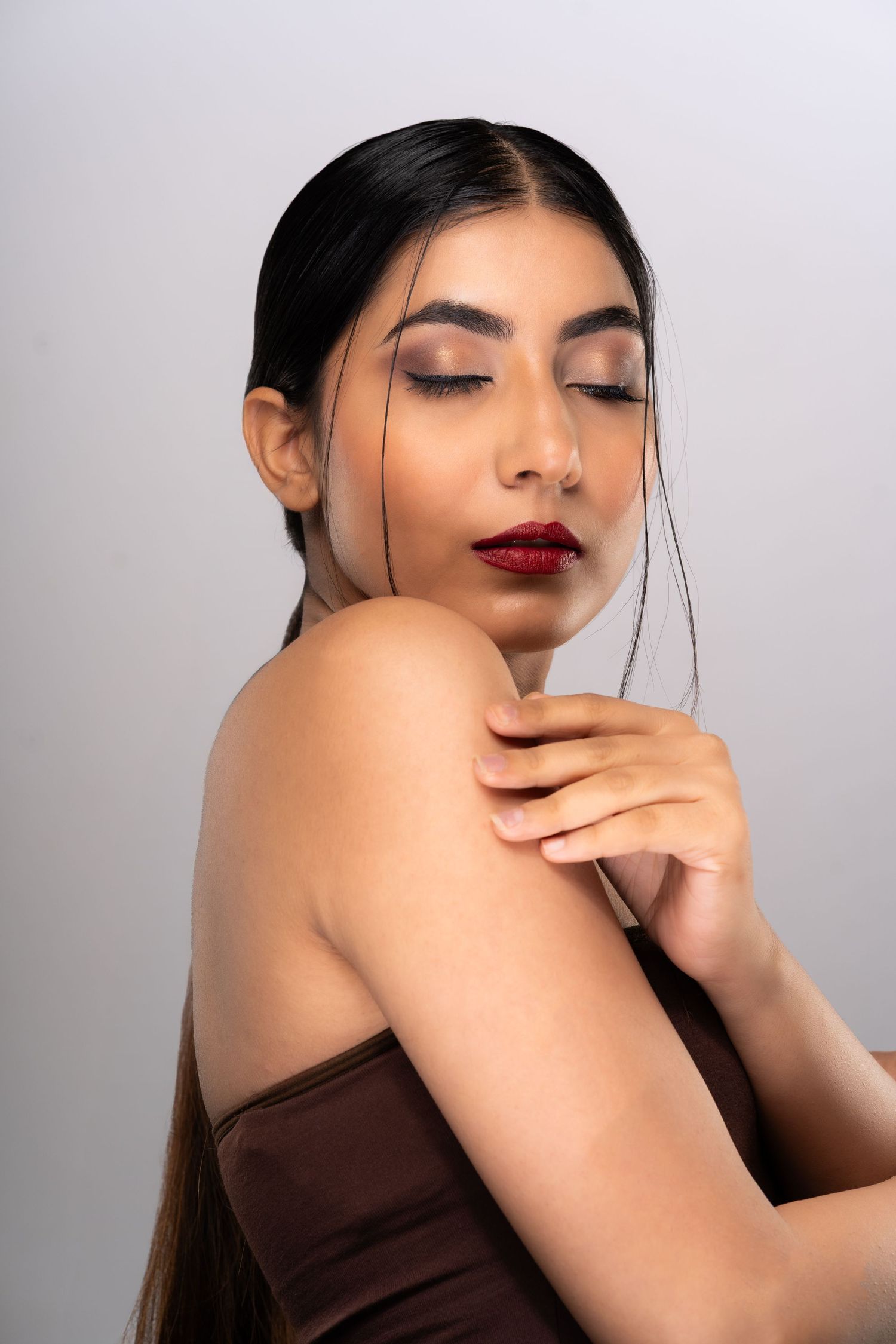 Young woman with red lips and dark hair poses gracefully with eyes closed, wearing an off-shoulder brown top.