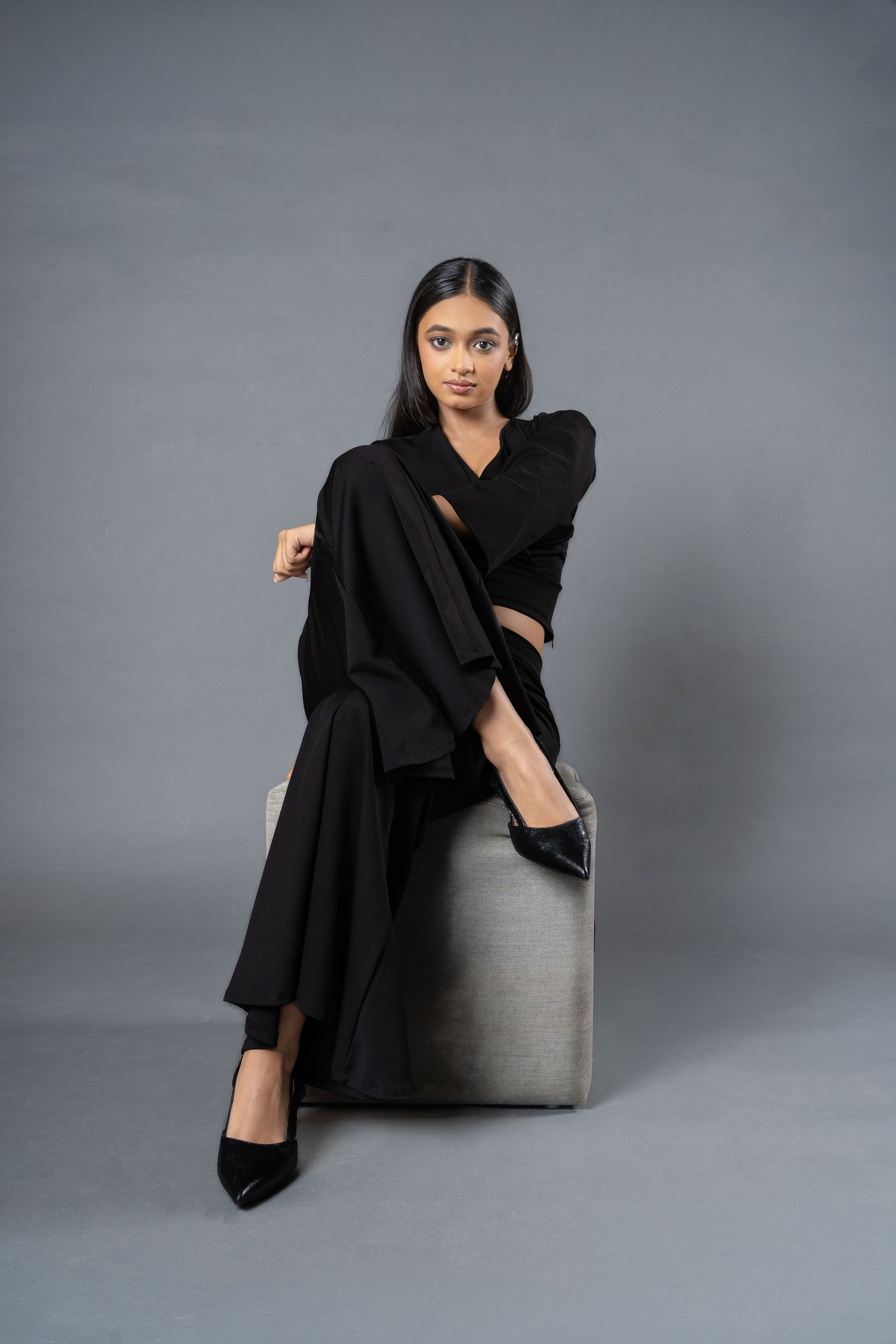 Young woman in black wide-leg pantsuit and heels posing on a gray cube stool.