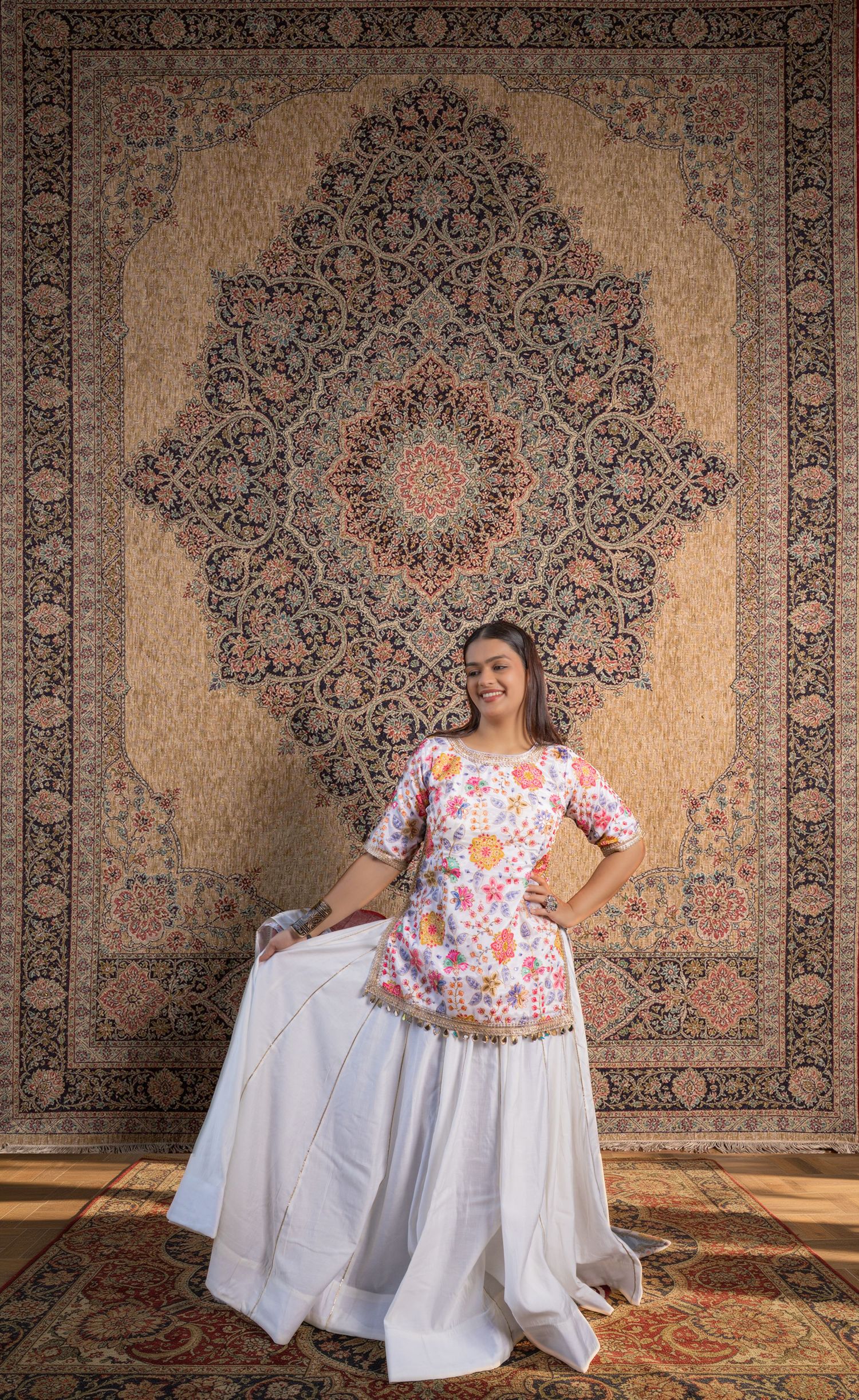 Woman in floral top and white skirt poses before ornate Persian carpet backdrop in elegant portrait.