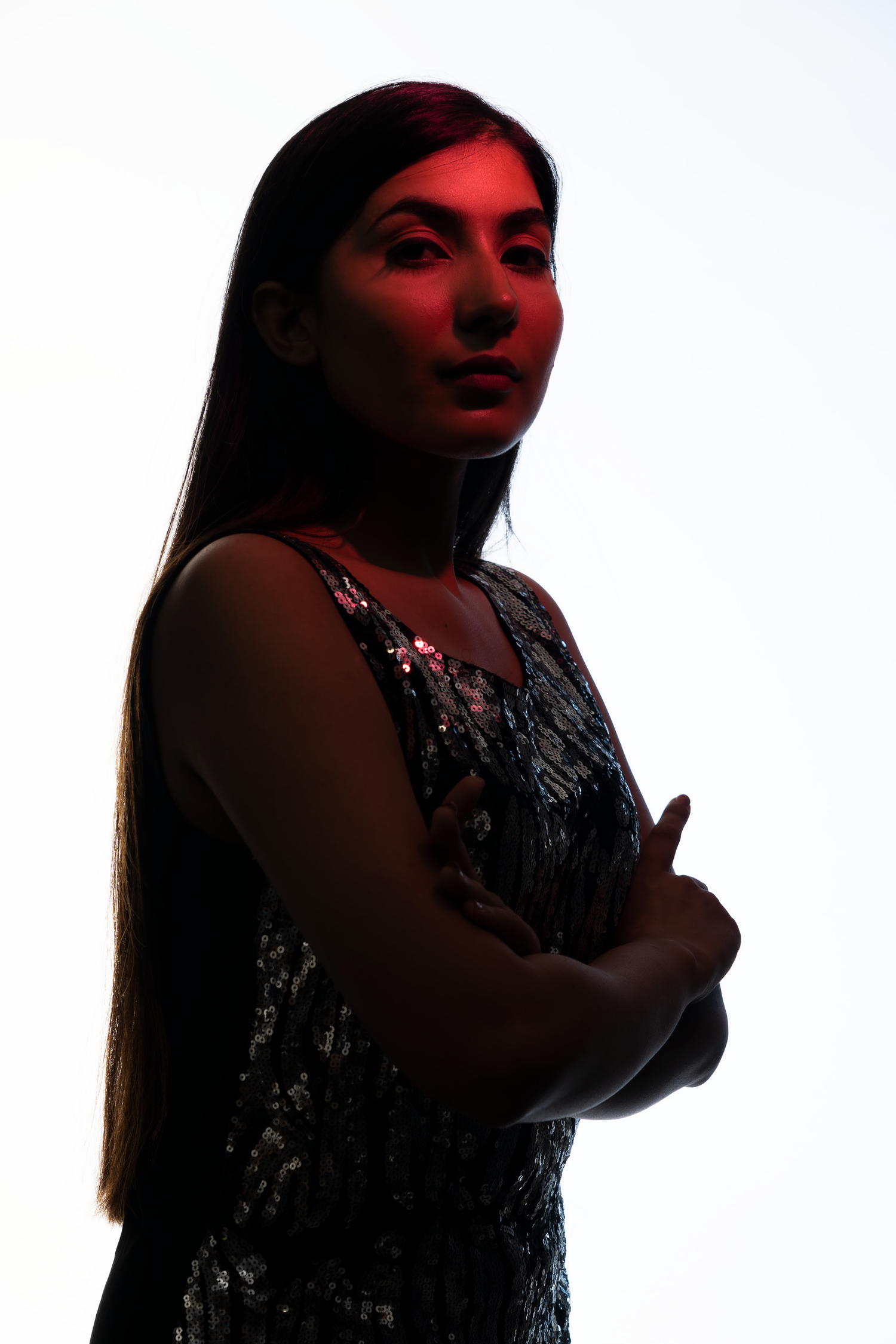 Young woman in sequin dress with arms crossed, dramatically lit with red light against white background.
