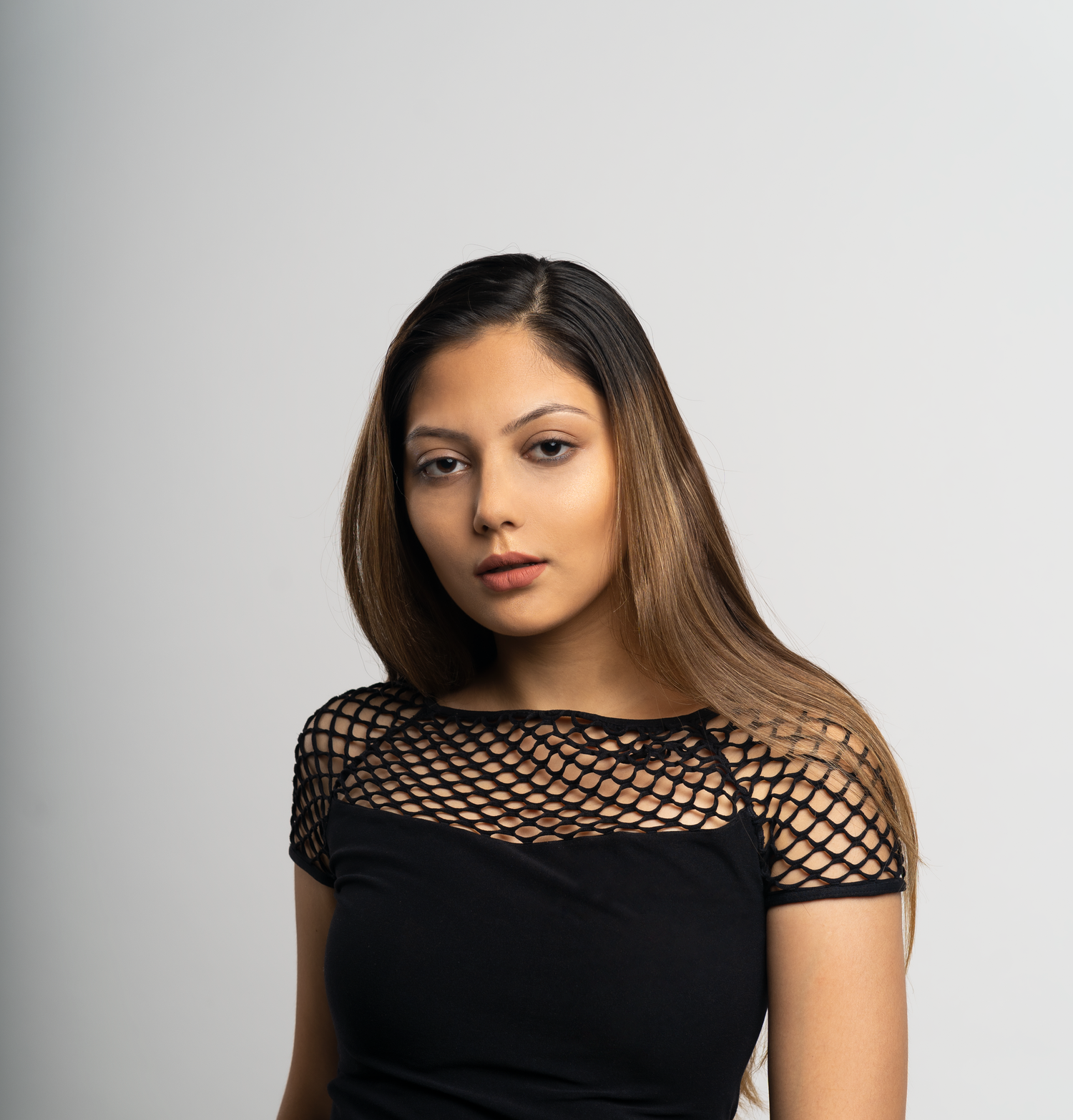 Young woman with long highlighted hair wearing a black mesh top, posing against a light gray background.