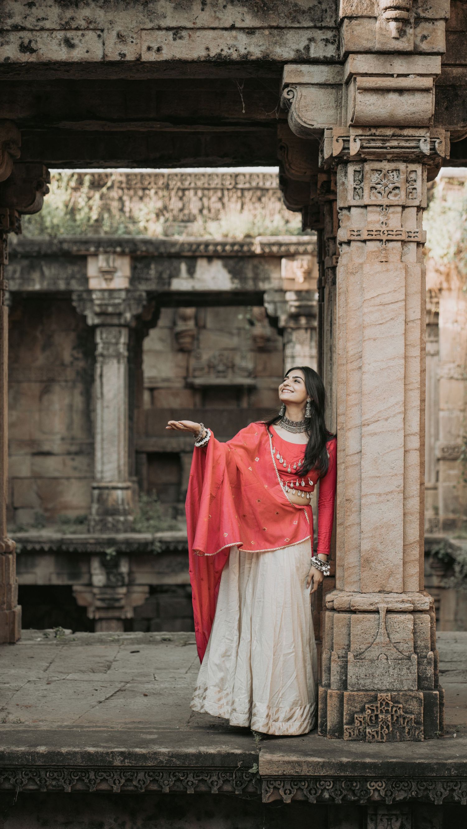 A woman in a red dupatta and white lehenga poses gracefully at an ancient Indian temple with ornate stone pillars.