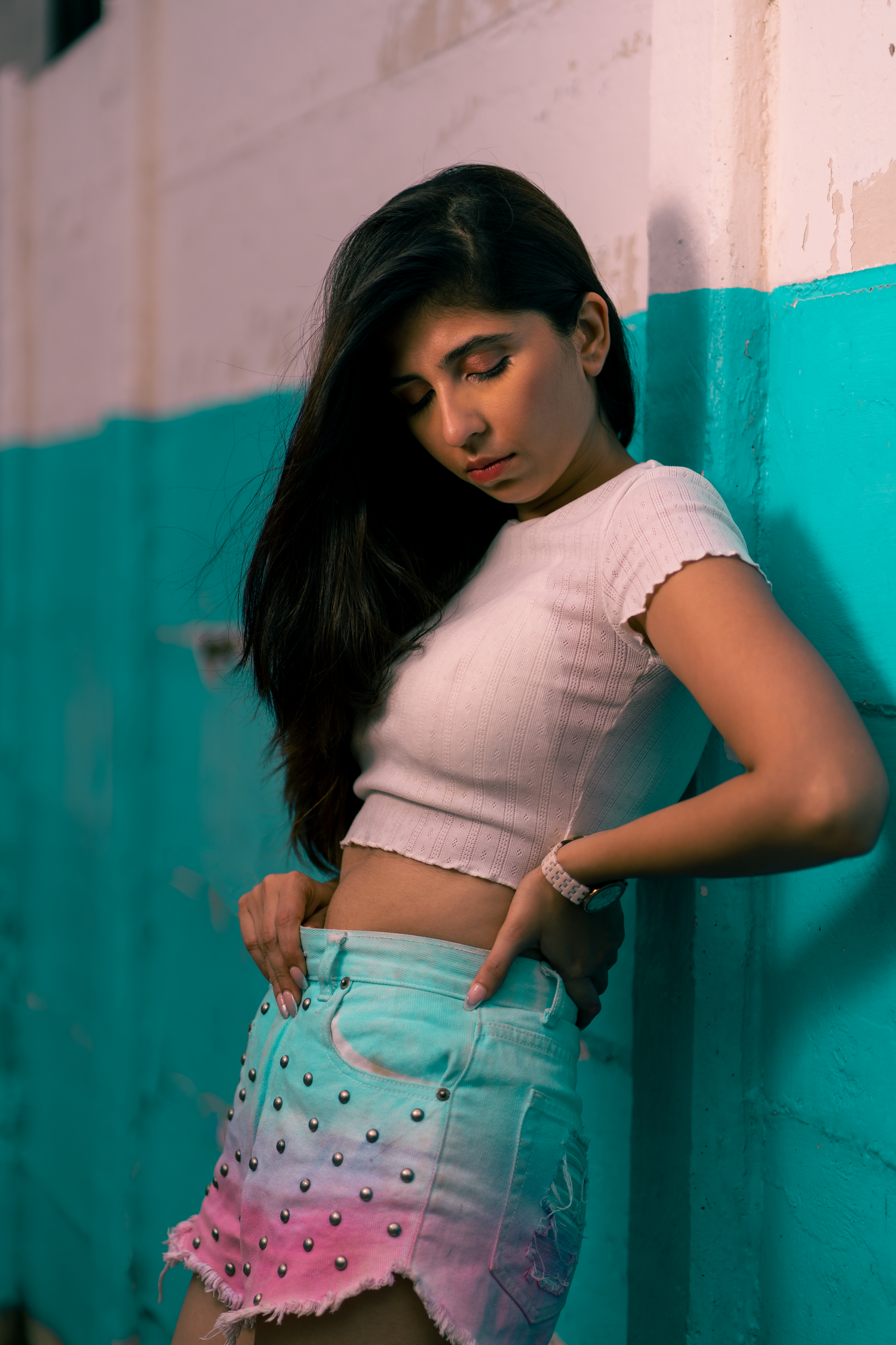 Young woman in white crop top and studded denim shorts poses against a teal and white wall.