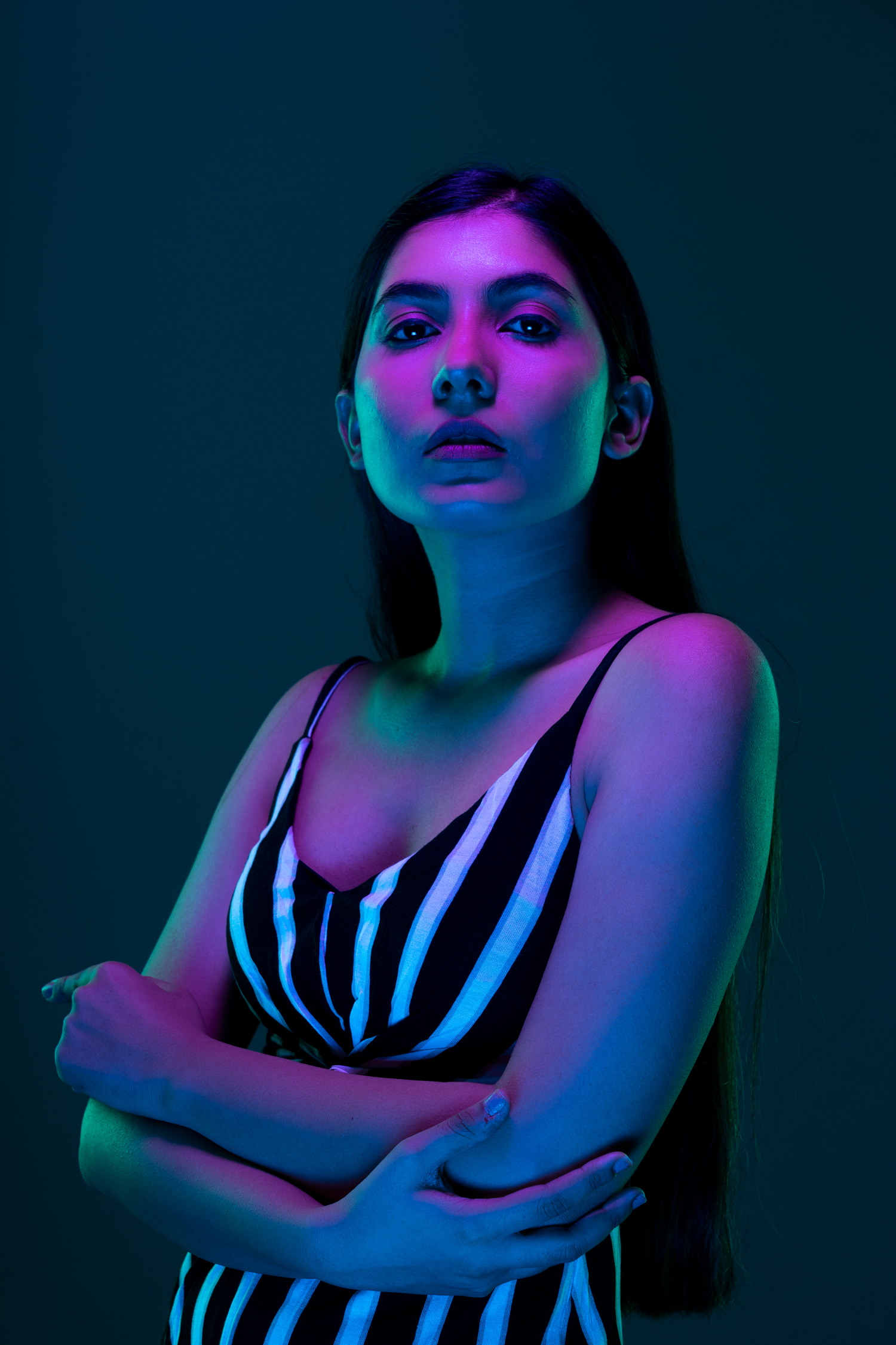 Young woman in striped dress poses with arms crossed under dramatic neon blue and pink studio lighting.