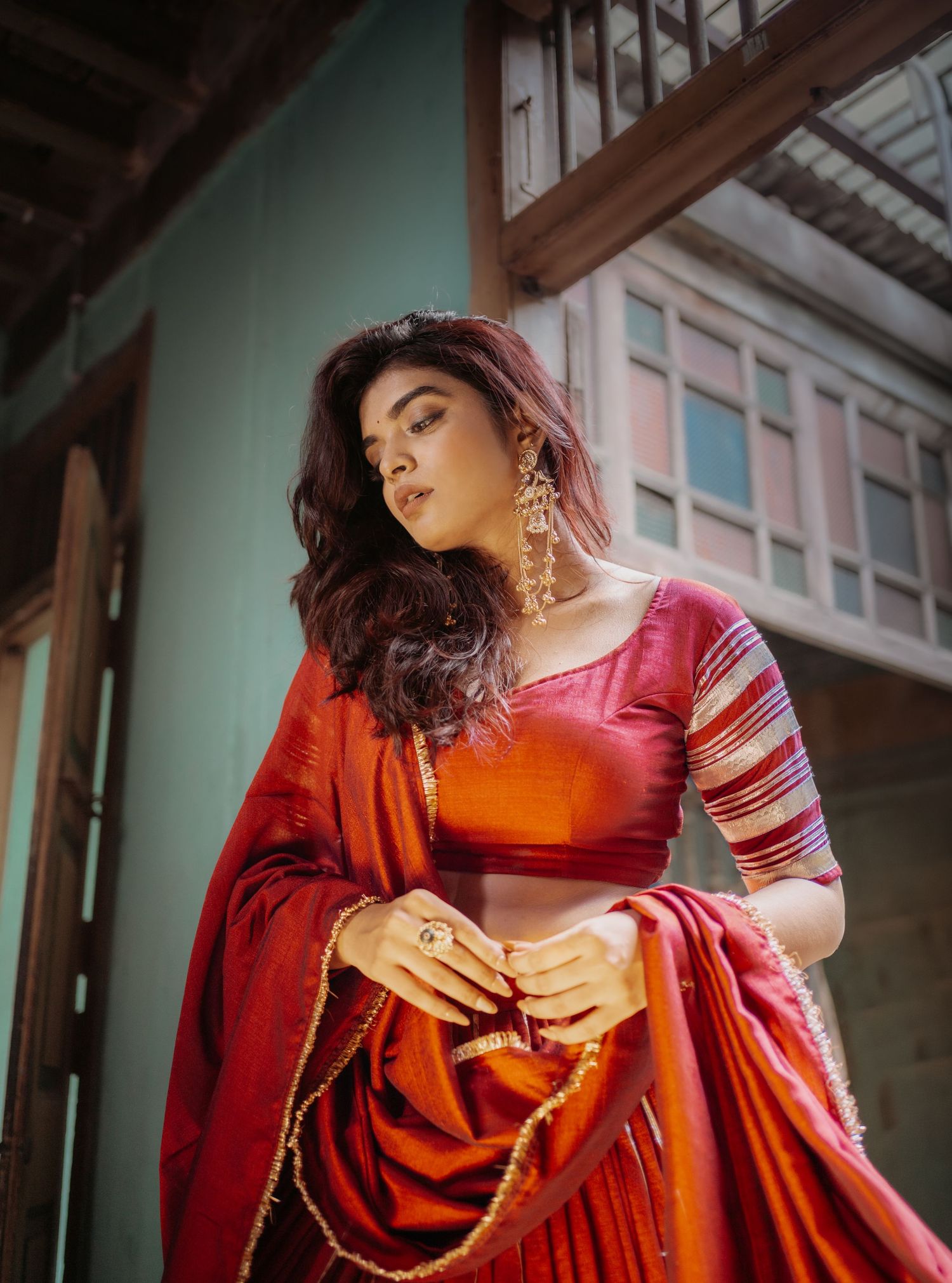 Woman in red and orange silk saree with gold trim and statement earrings poses gracefully outdoors.
