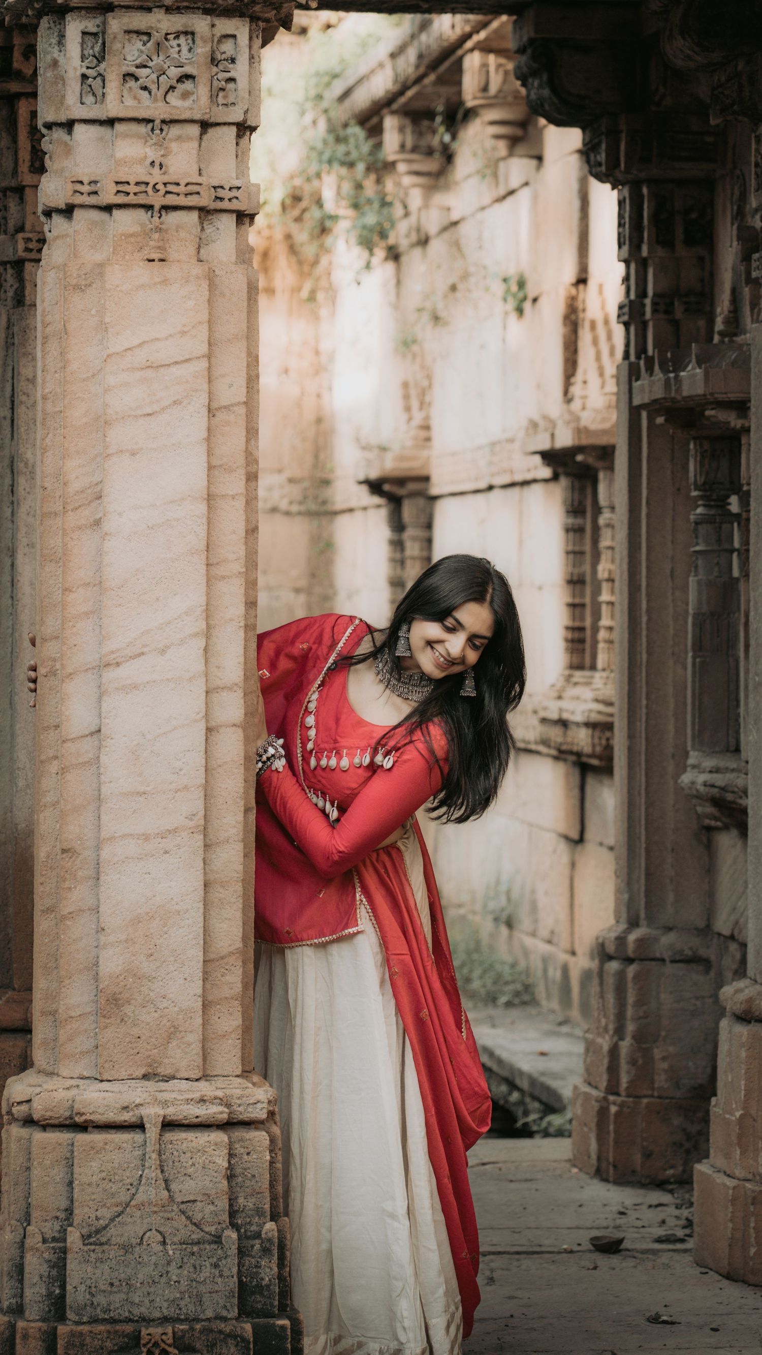 A young woman in a red and white traditional Indian outfit poses gracefully among ornate stone temple columns.