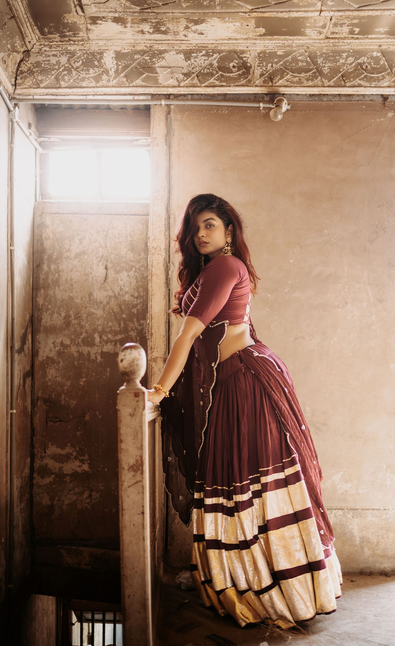 Woman in elegant maroon and gold lehenga poses on vintage staircase in rustic setting with dramatic lighting.