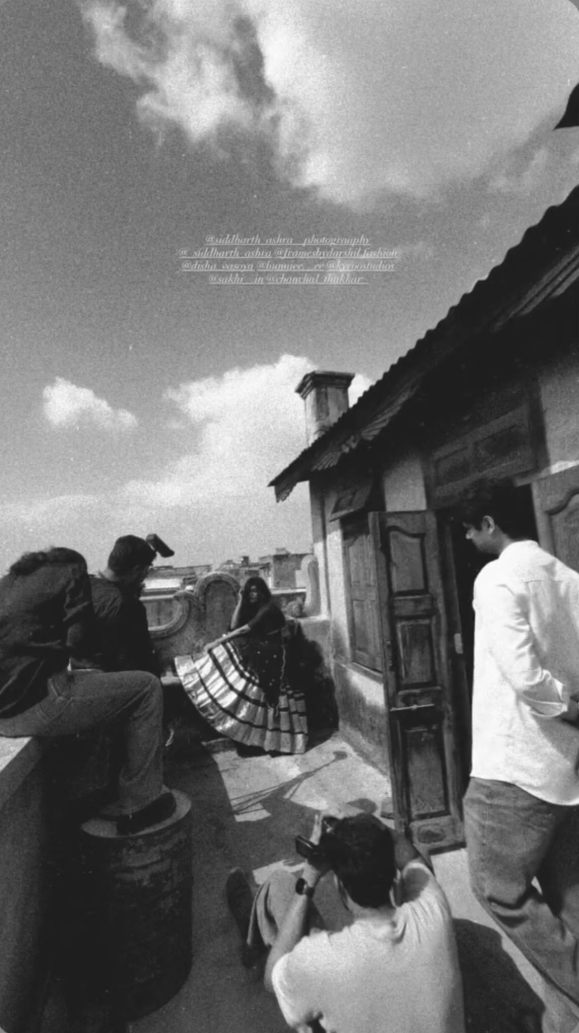 Black and white photo of a film shoot on a rooftop with crew members and a woman in a traditional outfit posing.
