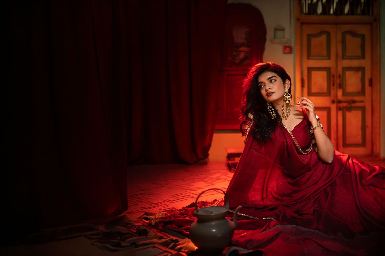 Woman in elegant red saree poses dramatically on floor bathed in moody red light with vintage decor.