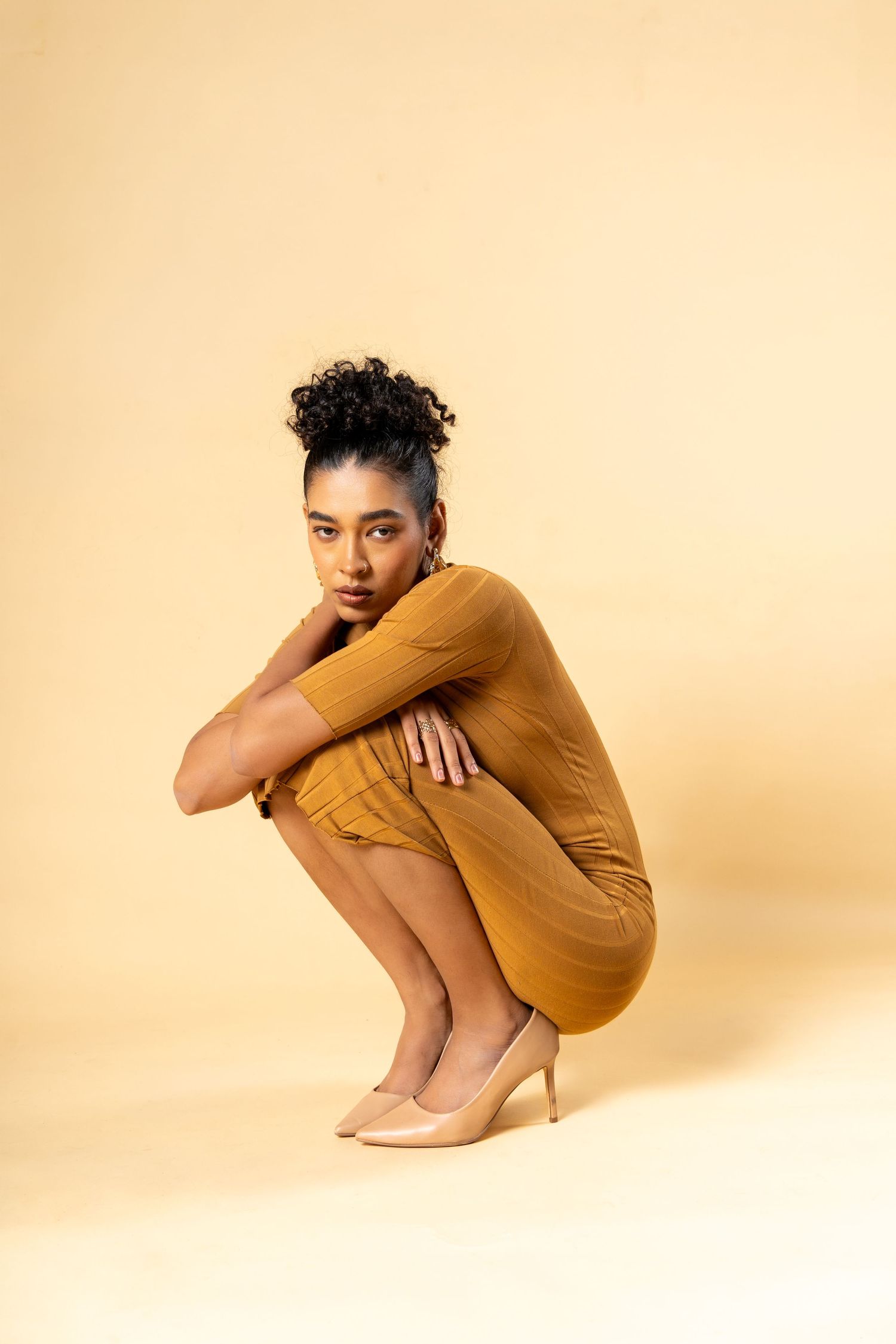 A woman in a mustard yellow dress and nude heels crouches gracefully against a beige background.