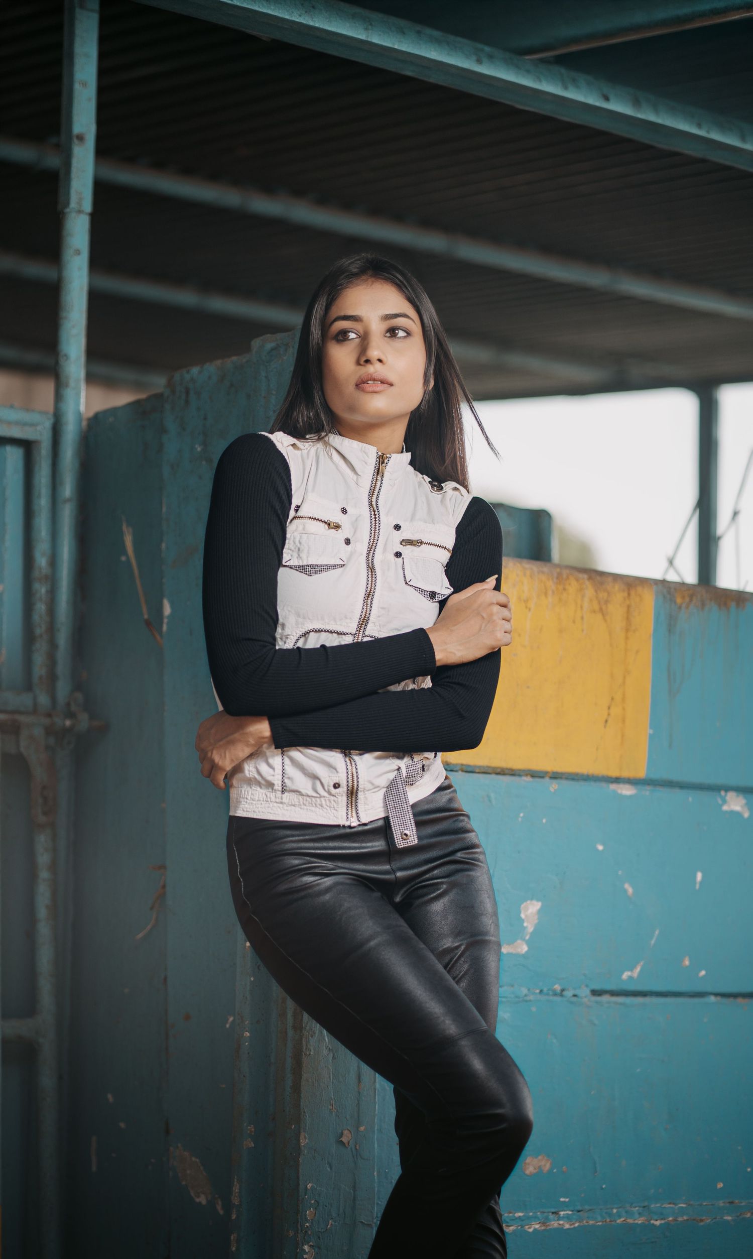 Young woman in white vest and black leather pants leans against teal wall in urban setting.