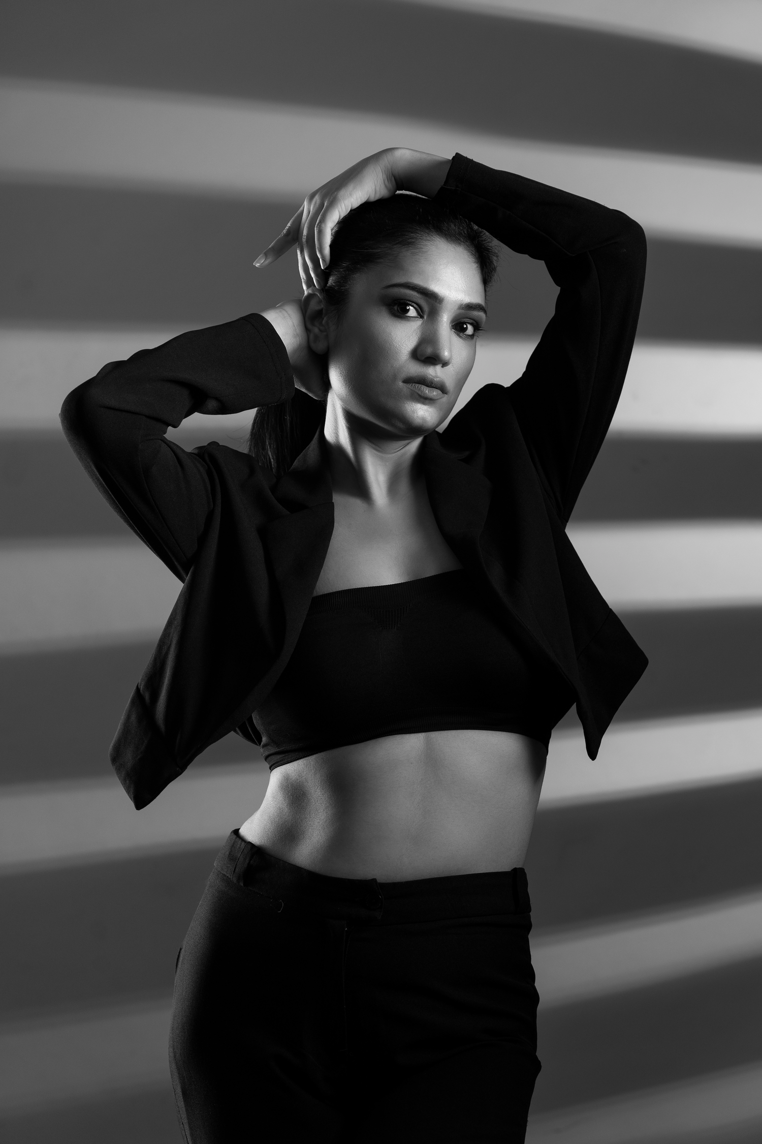 Black and white photo of a fit woman in crop top and blazer posing with hands on head against striped background.