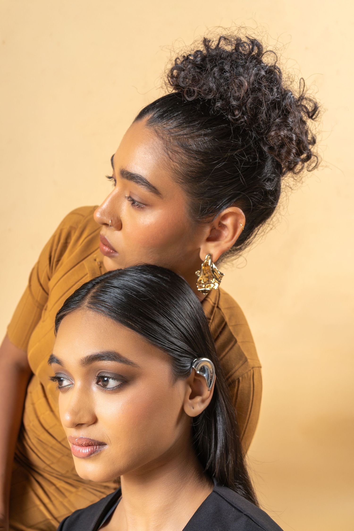 Two women pose together, one with curly updo and gold earring, other with sleek dark hair and hearing aid, against beige background.