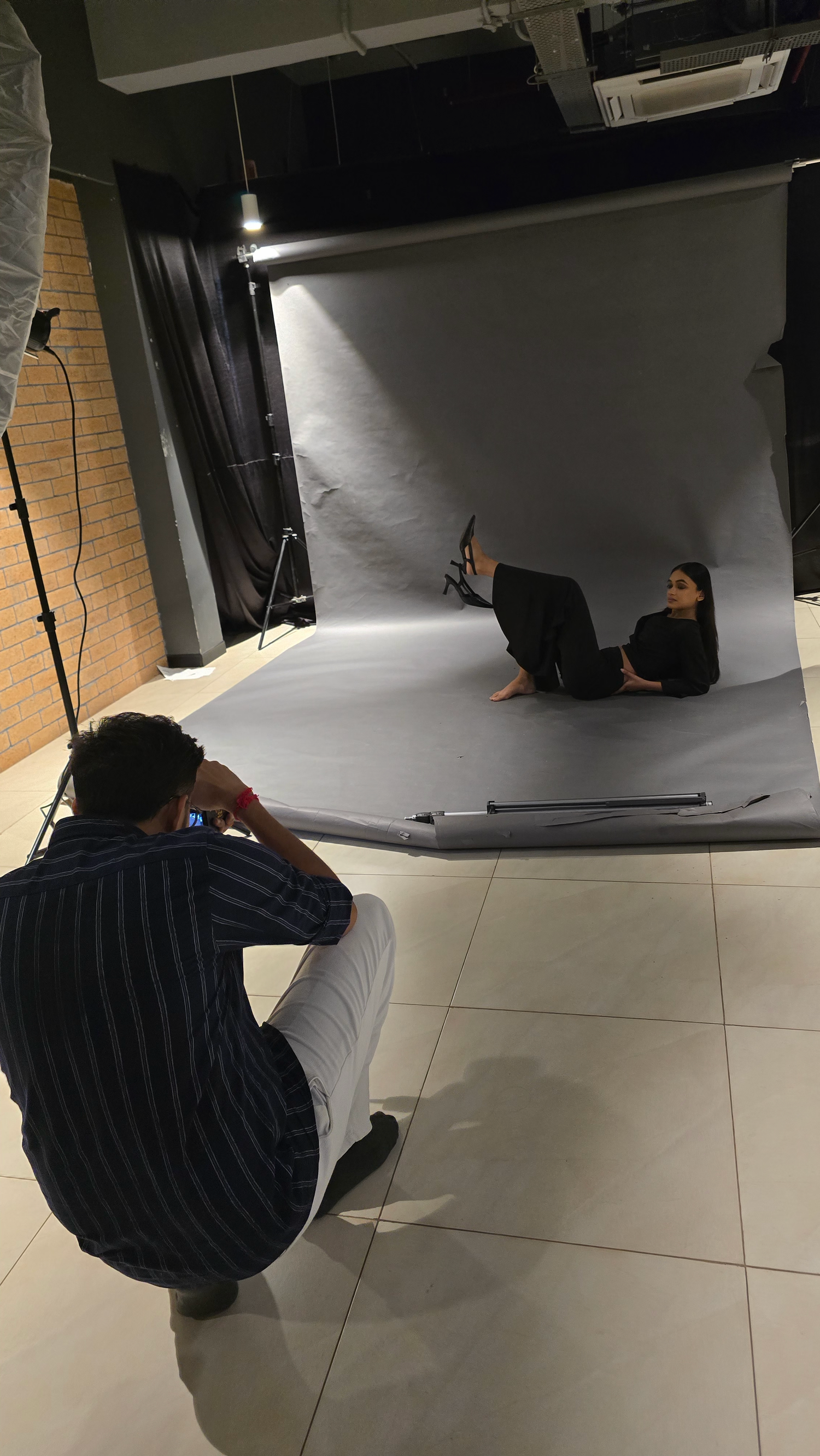Photographer crouching on studio floor capturing a model in black outfit posing on gray backdrop with professional lighting setup.