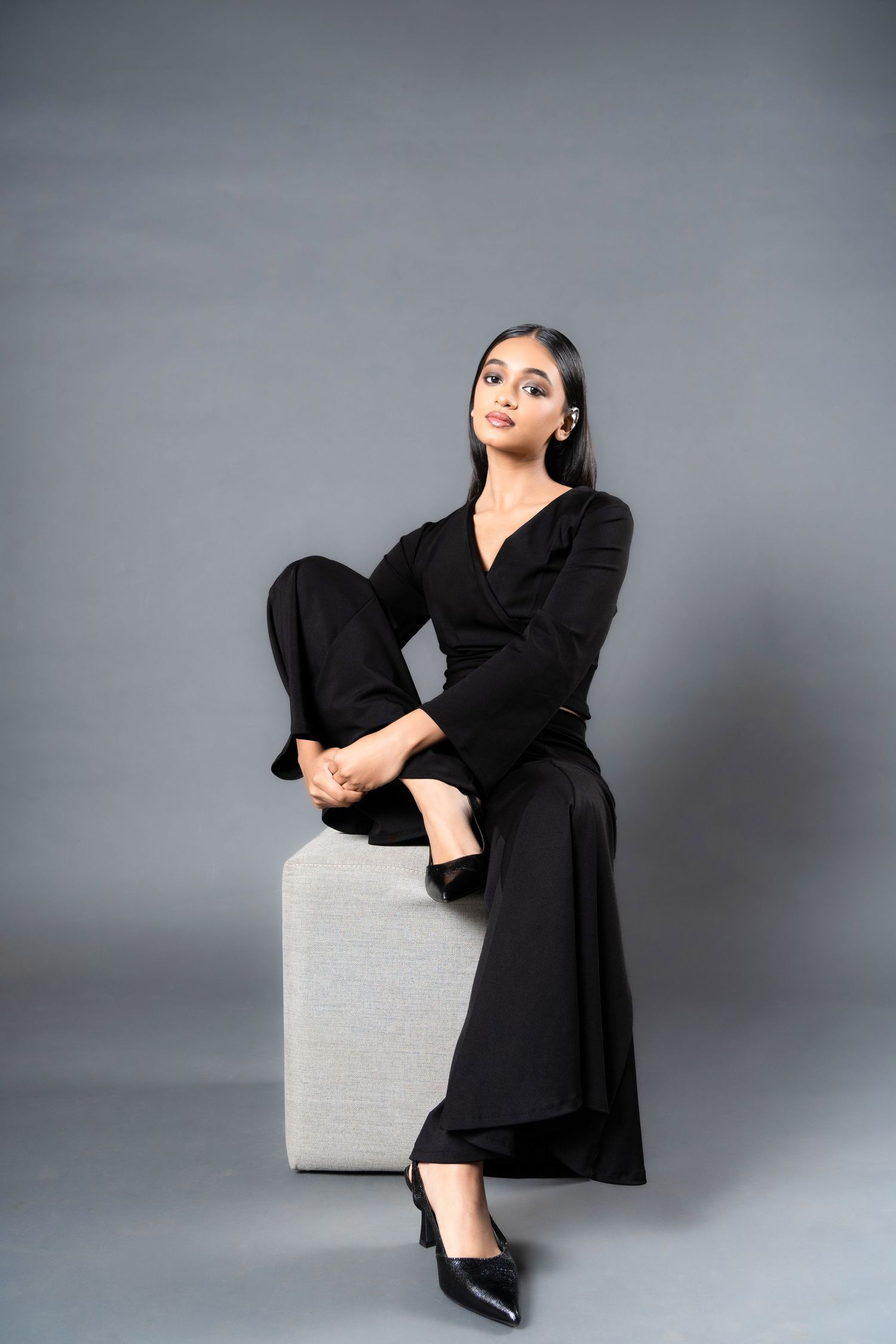 Woman in black jumpsuit and heels seated on a gray cube against a neutral studio background.
