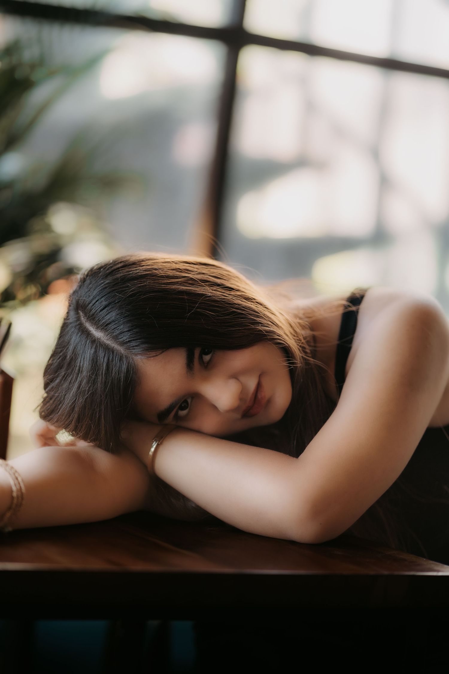 Young woman with dark hair rests her head on crossed arms on a wooden table, gazing softly at the camera.