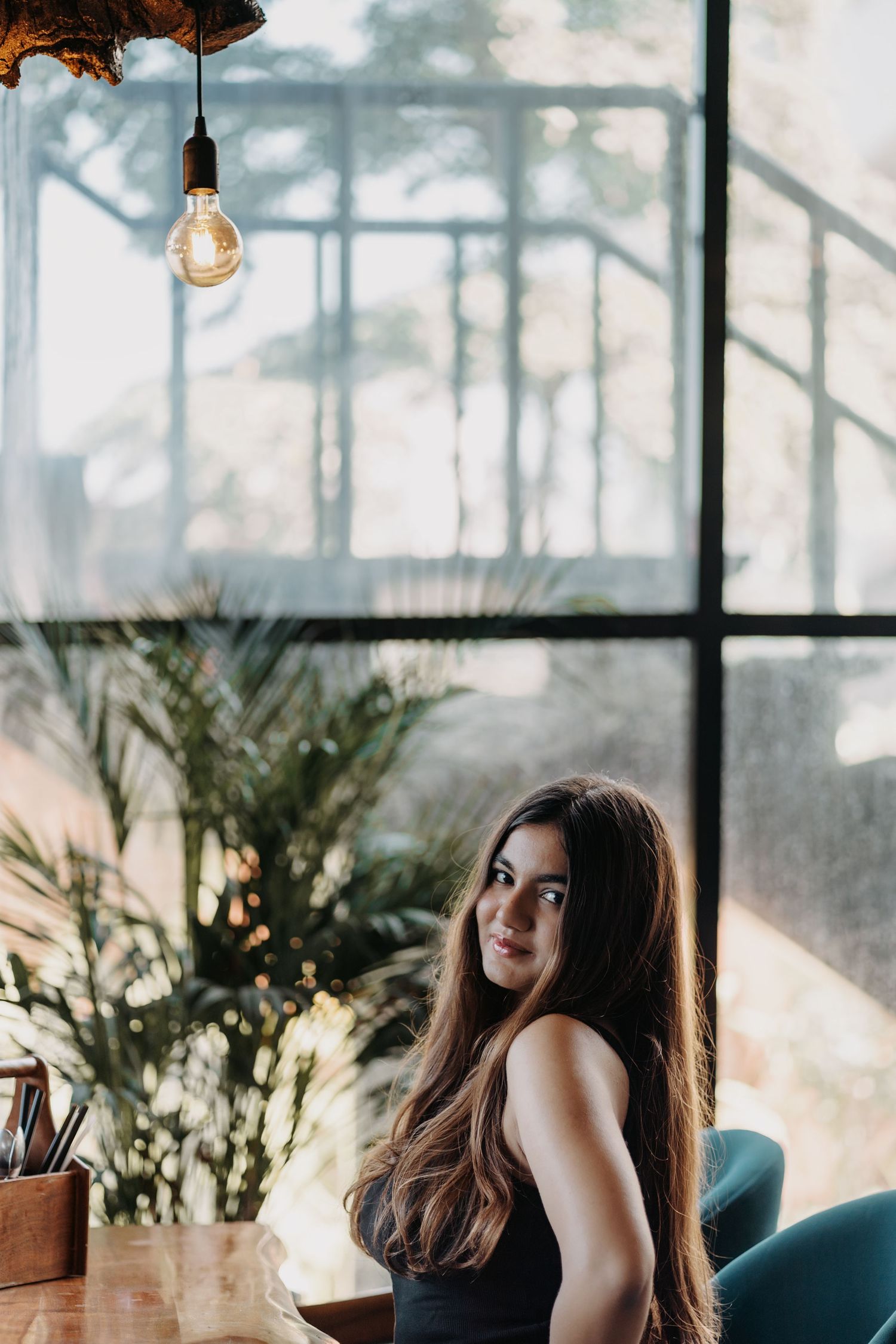 Young woman with long hair smiling over shoulder in stylish cafe with Edison bulb and large windows.