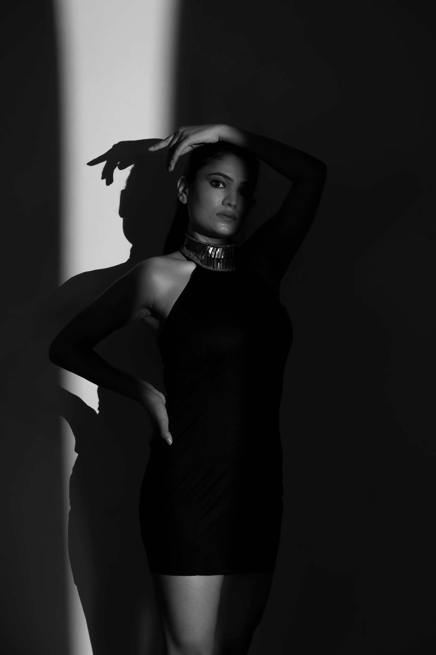 Black and white photo of a woman in a black mini dress and choker necklace posing dramatically with arms raised in moody lighting.