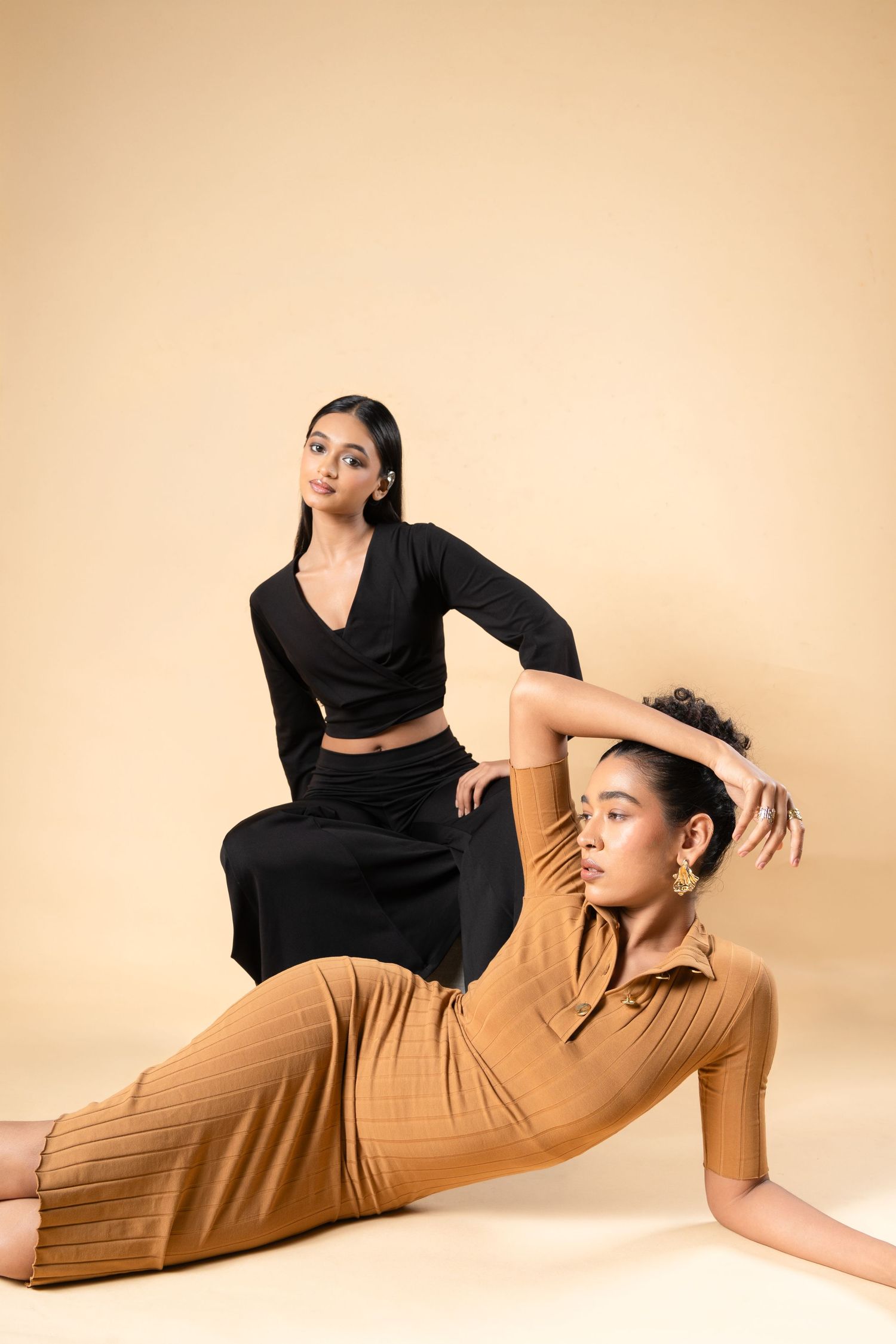 Two models pose elegantly, one in black crop set, other in tan ribbed dress, against beige background.