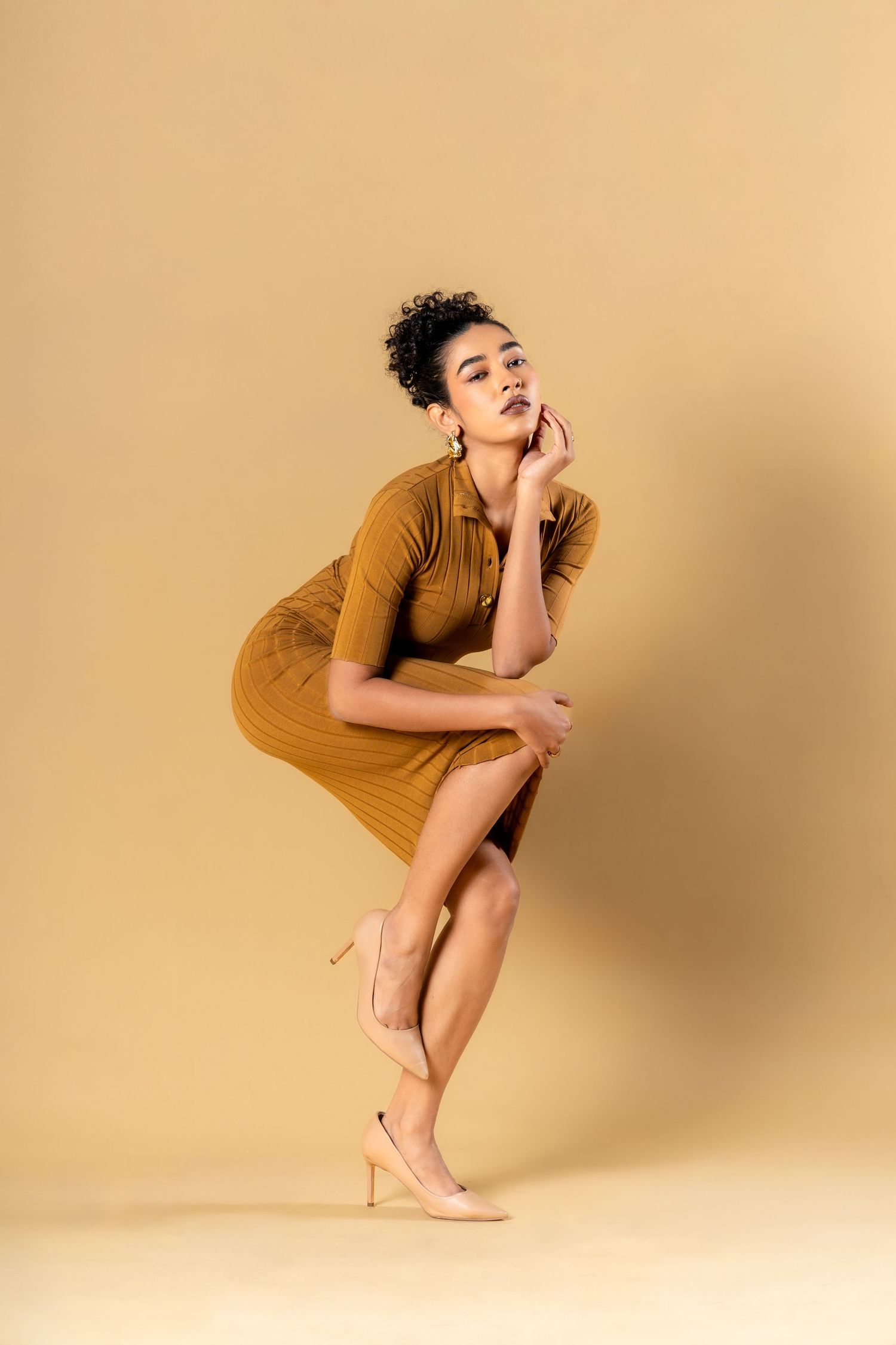 Woman in brown ribbed dress and nude heels poses gracefully against a beige background.