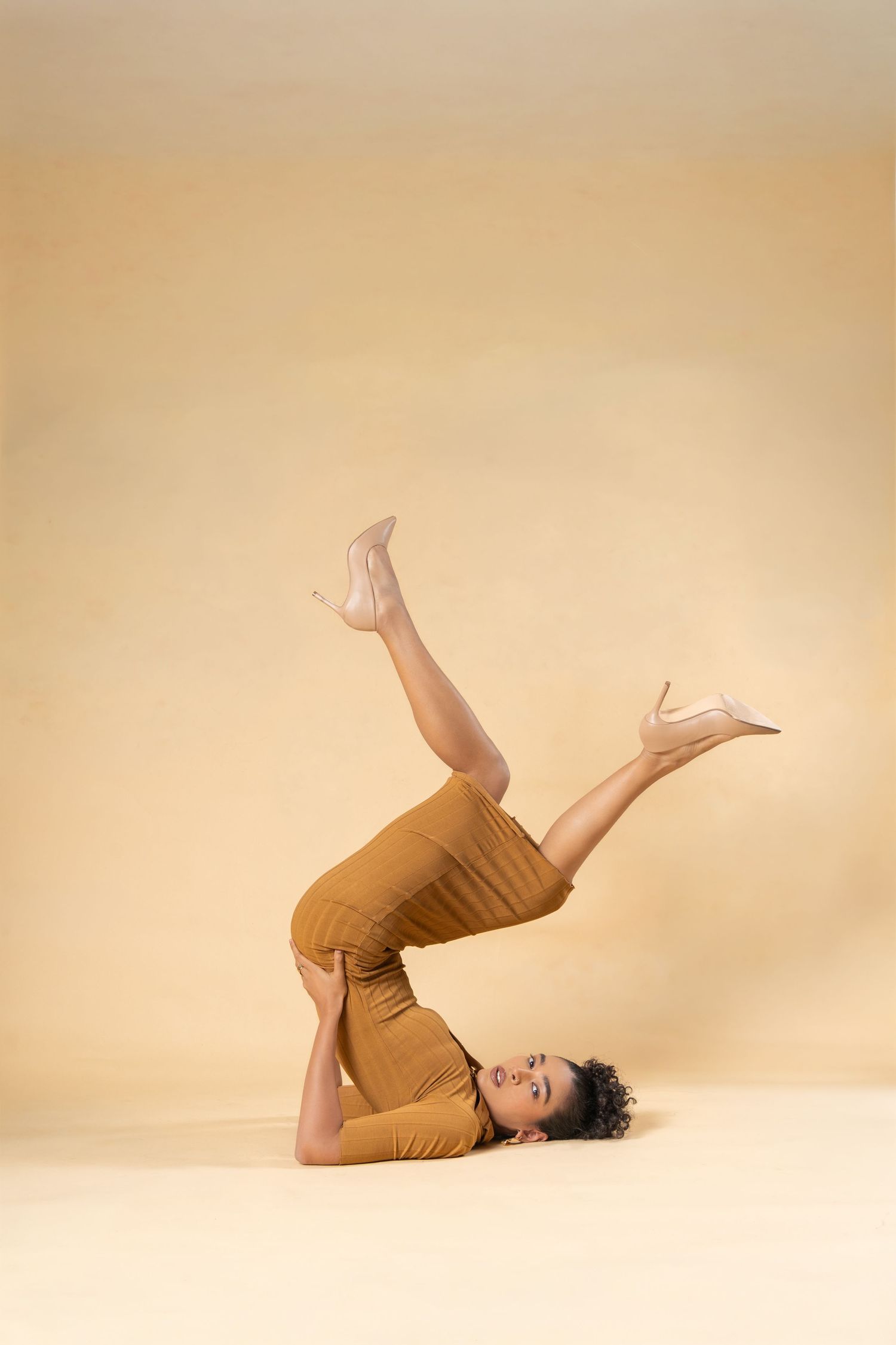 Woman in tan dress and heels performing a shoulder stand pose against a beige studio background.