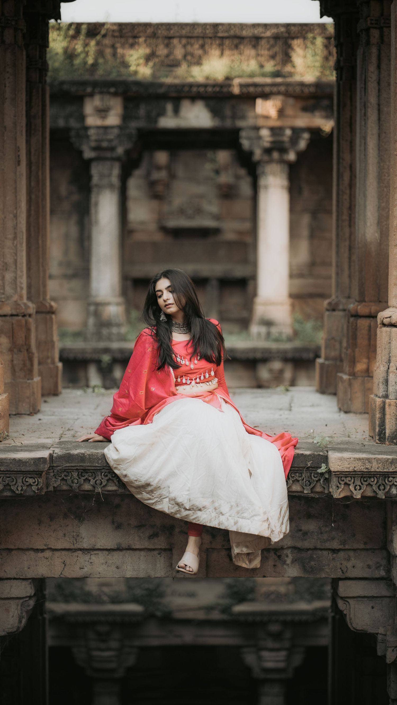 Woman in red kurta and white skirt posing gracefully at historic Indian stone architecture.