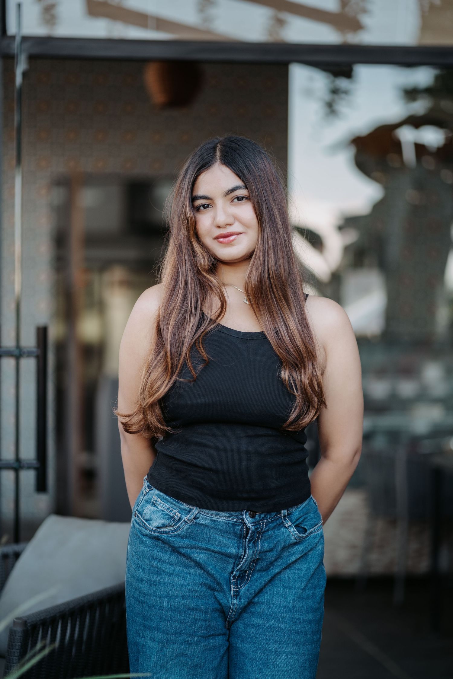 Young woman with long brown hair wearing a black tank top and blue jeans standing outside a modern building.