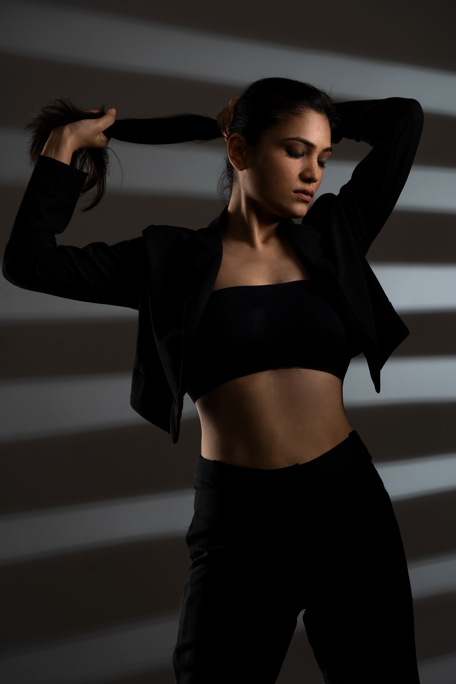 Woman in black crop top, blazer, and pants poses dramatically against striped shadow wall backdrop.