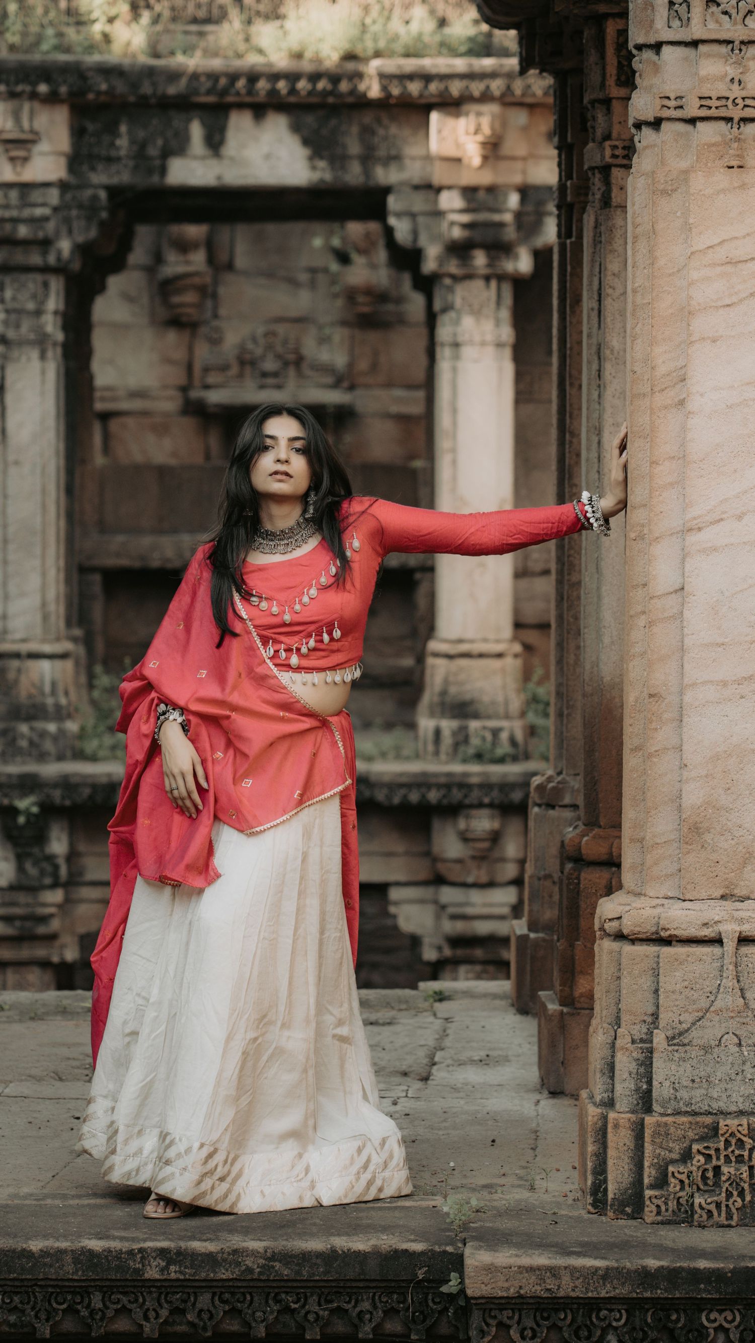 A woman in a red embroidered top and white lehenga poses gracefully at an ancient stone temple.