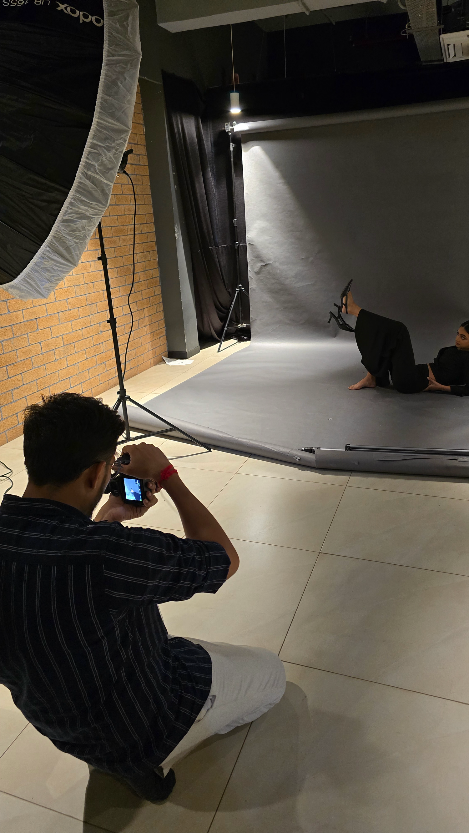 Photographer kneeling with camera in professional photo studio with gray backdrop and studio lighting equipment.