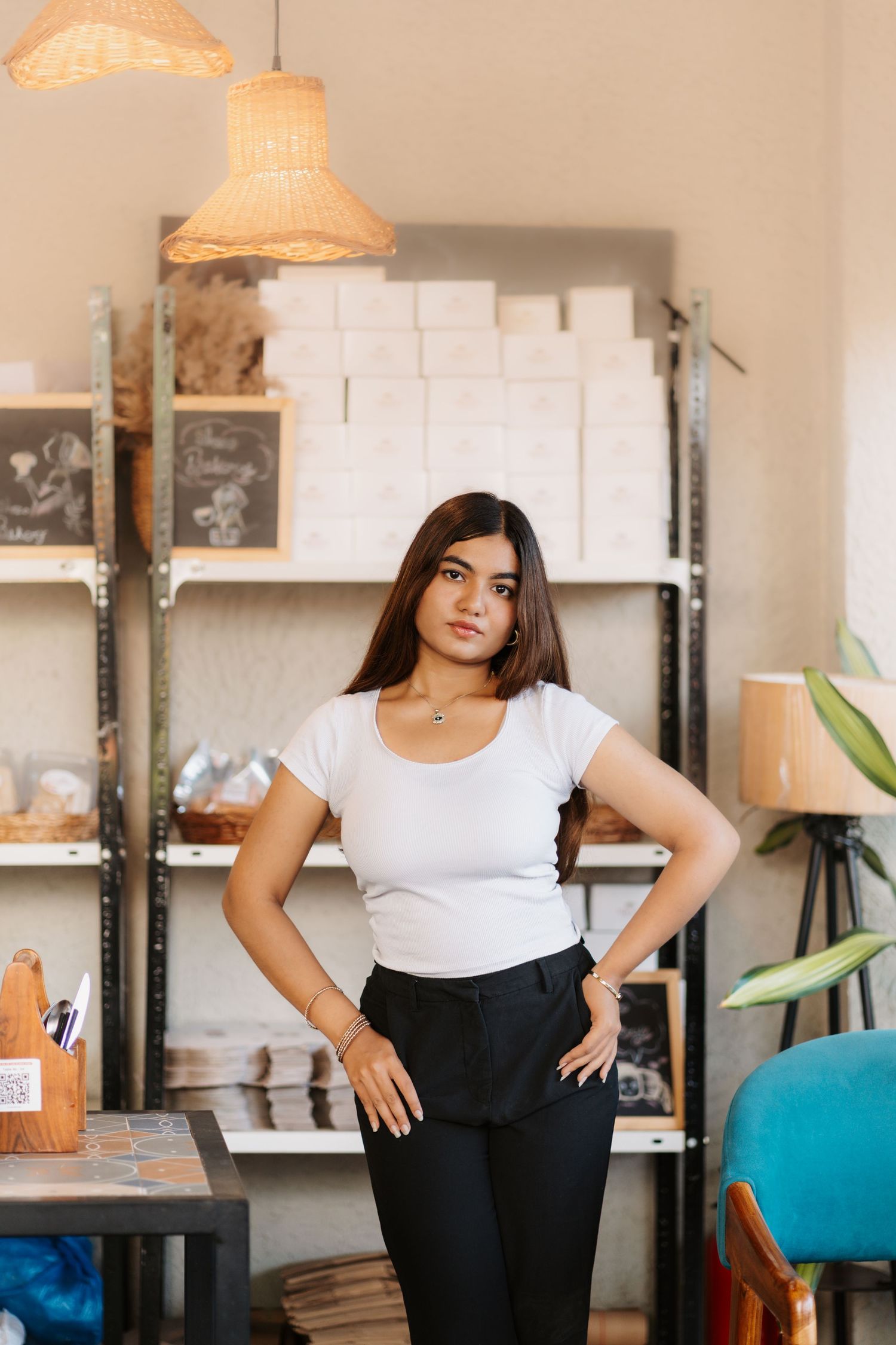 Young woman in white t-shirt and black pants poses confidently in a cozy artisan shop with shelves and warm lighting.