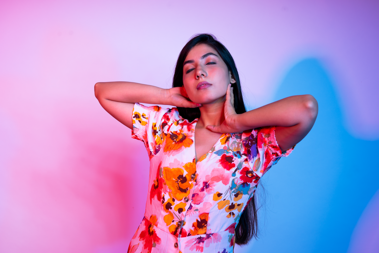 Woman in floral dress poses against pink and blue neon lit background with hands behind neck.