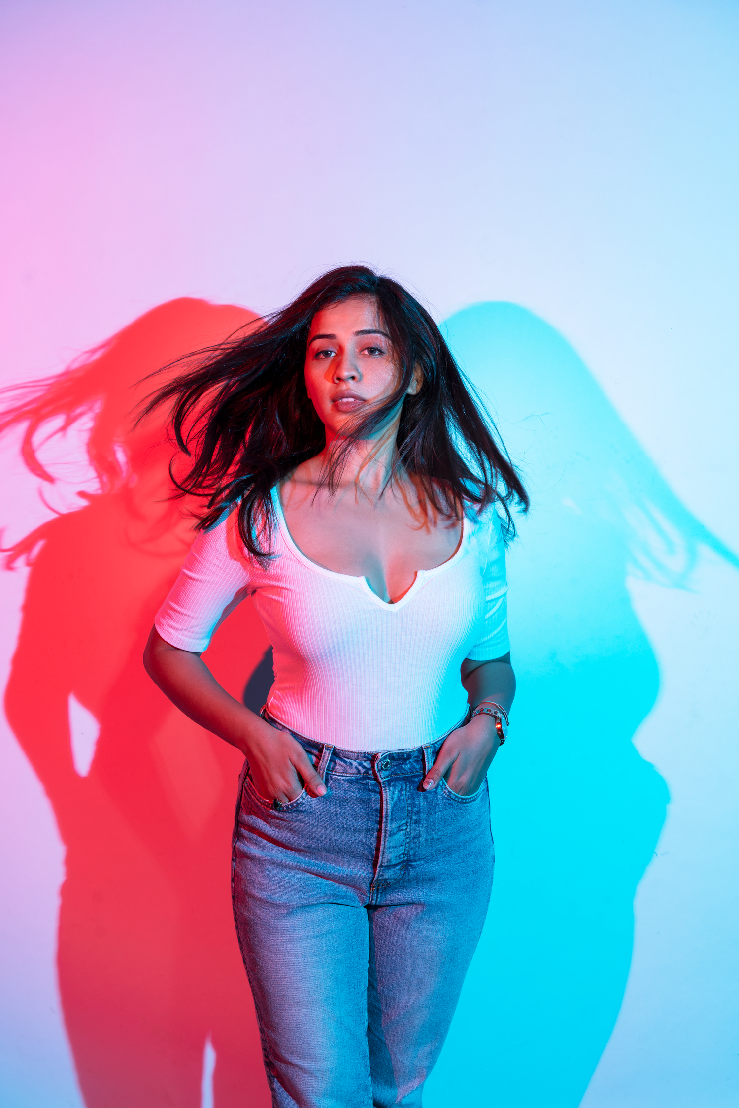 Young woman in white bodysuit and jeans poses against colorful pink and blue neon lit background with dramatic shadows.