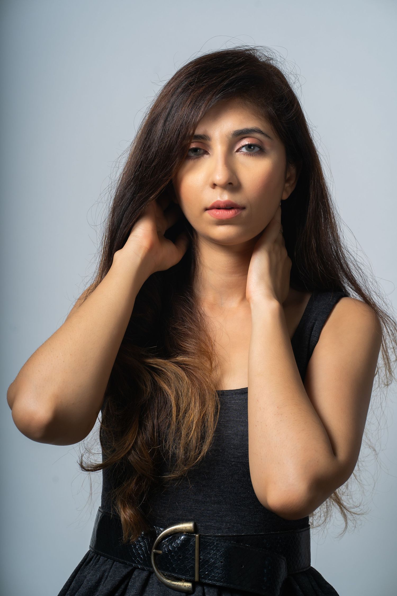 Young woman with long dark hair in black dress and belt, posing with hands near neck against gray background.