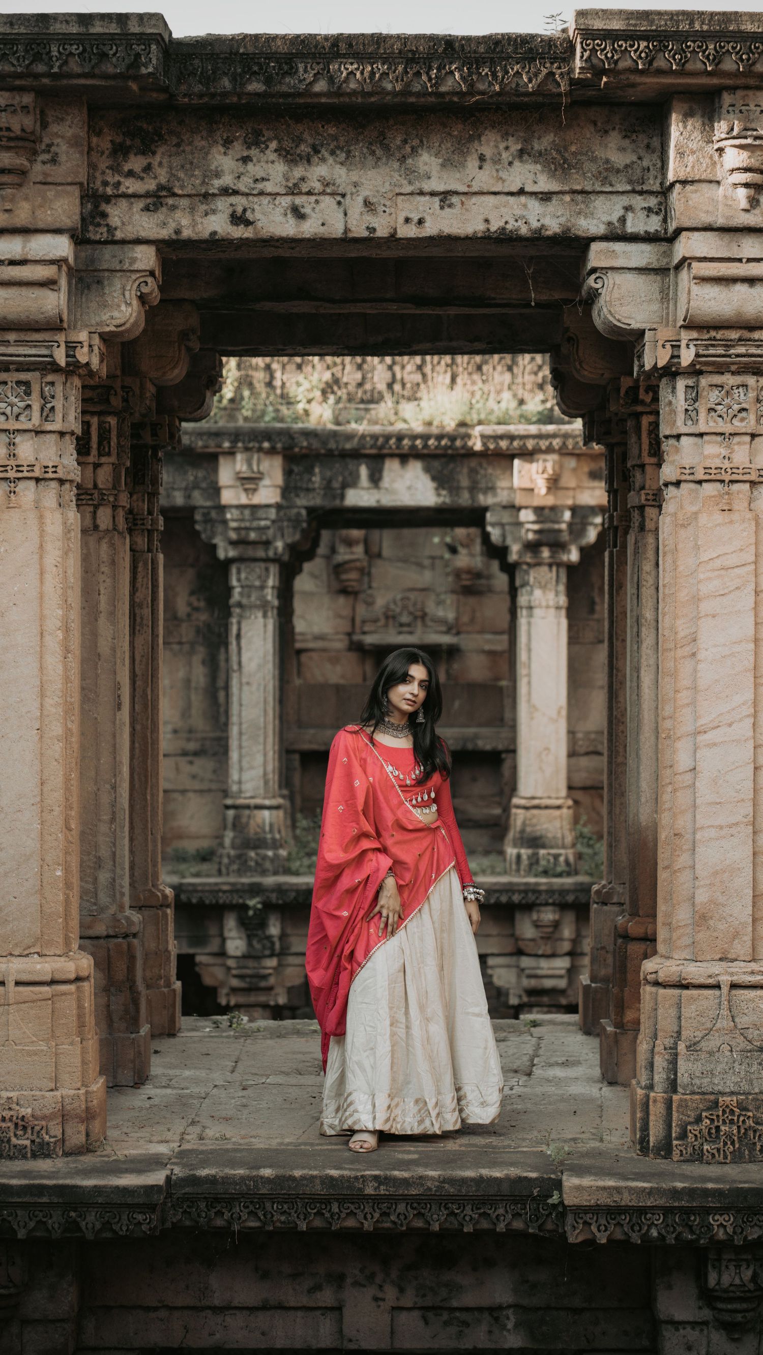 Woman in red and white traditional Indian attire standing in ancient stone temple archway.
