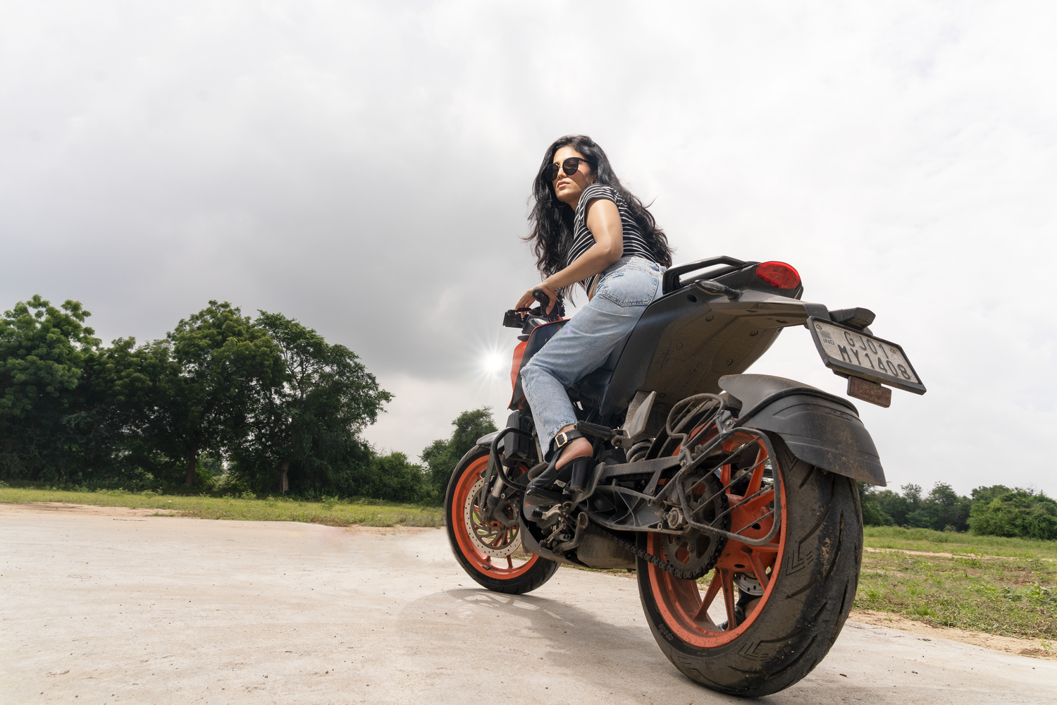 Woman in sunglasses posing on an orange and black KTM motorcycle on an open road with cloudy skies.
