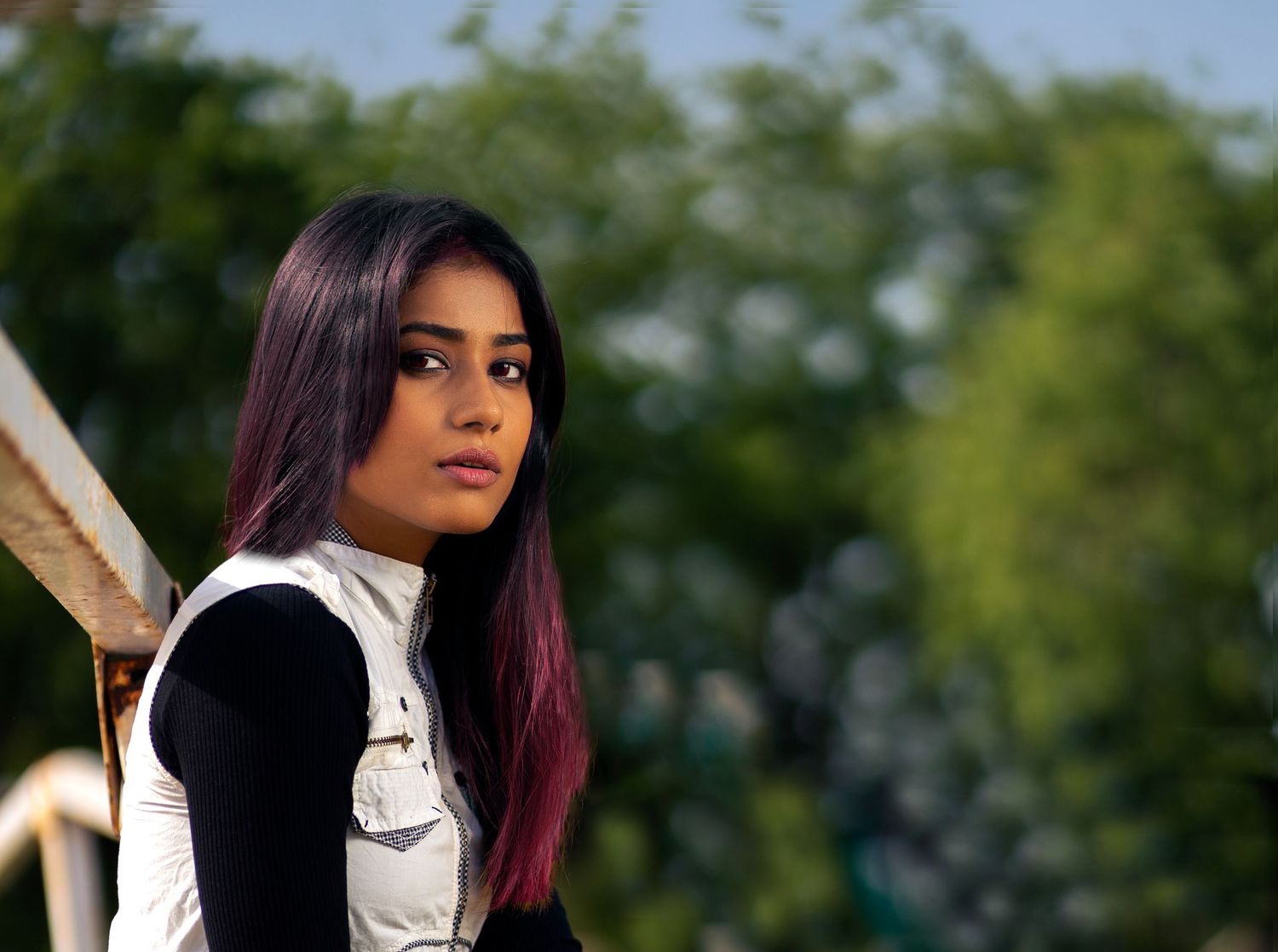 Young woman with long dark hair and purple highlights poses outdoors with green trees in the background.