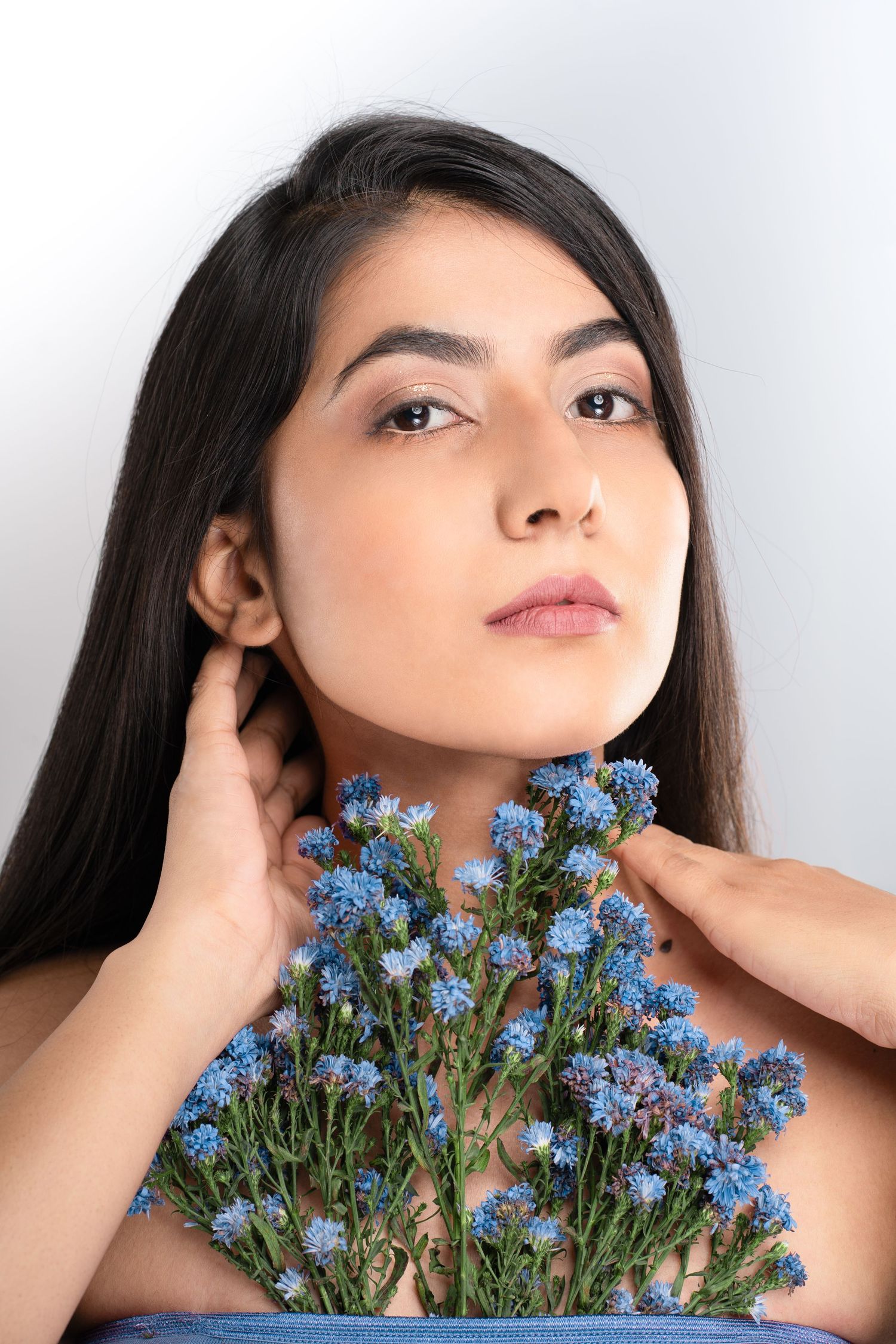 Young woman with long dark hair holds blue wildflowers near her neck against a light gray background.