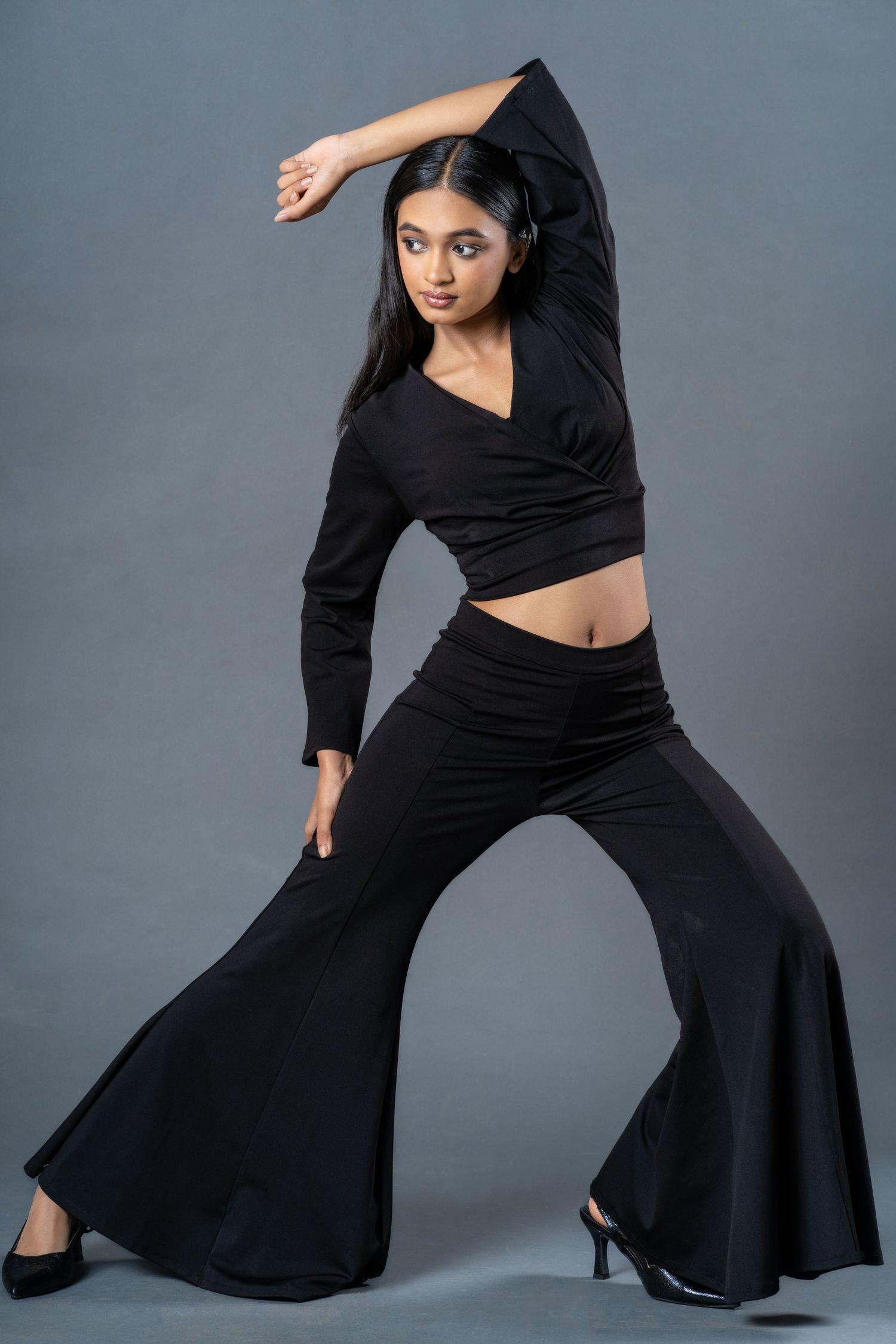 Young woman in black crop top and flared pants striking a dance pose against gray background.