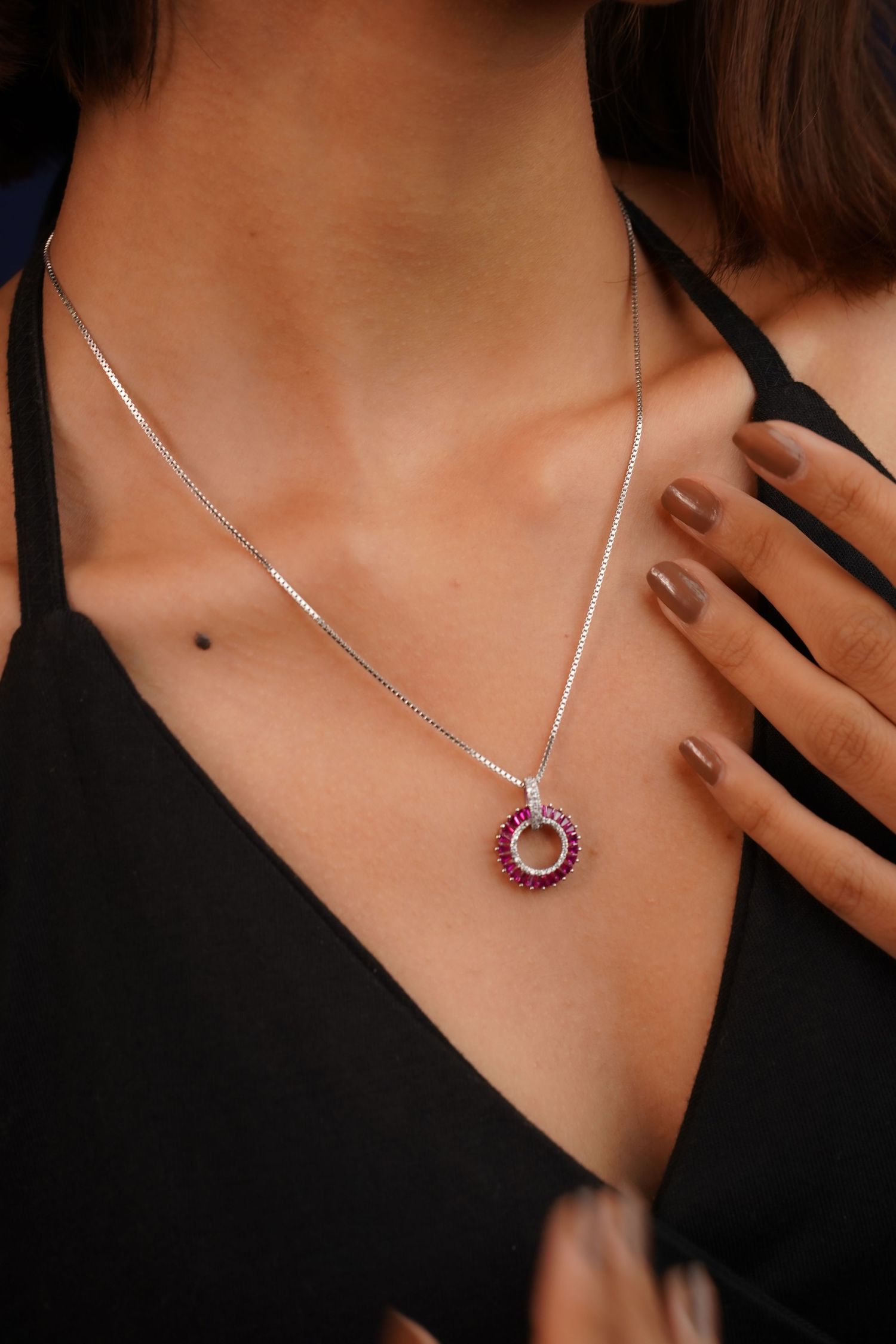 Woman wearing a silver chain necklace with a circular ruby and diamond pendant over a black top.