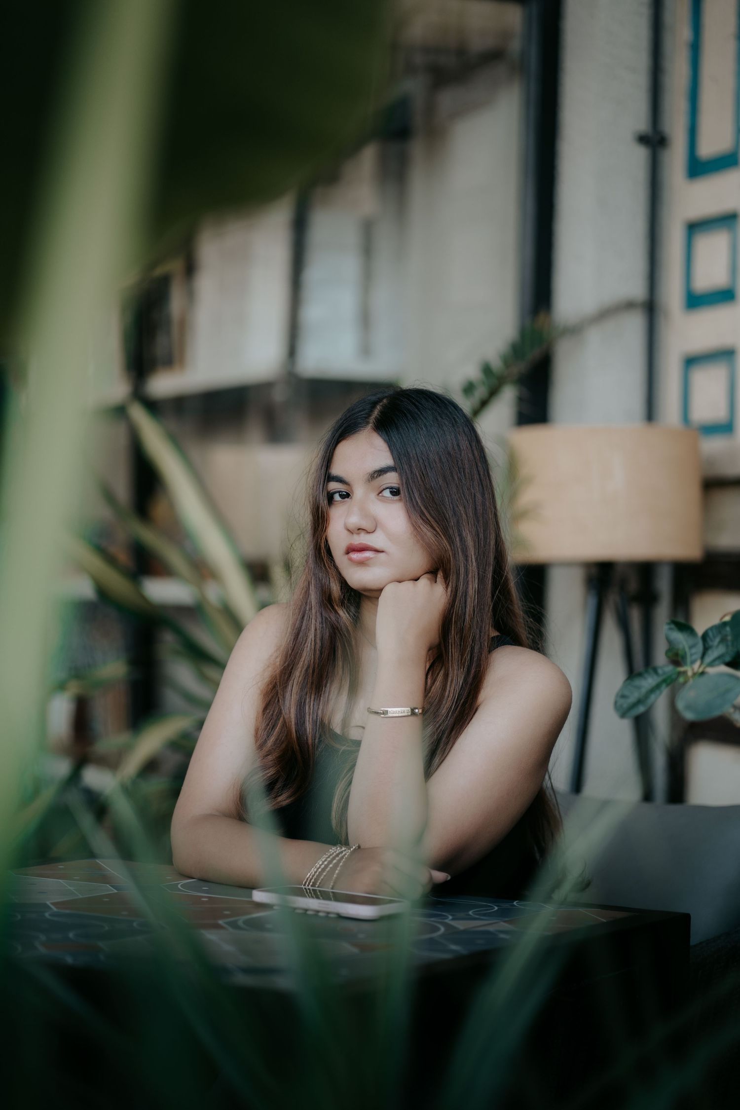 Young Asian woman with long hair sits thoughtfully at a cafe table, resting chin on hand, surrounded by lush green plants.