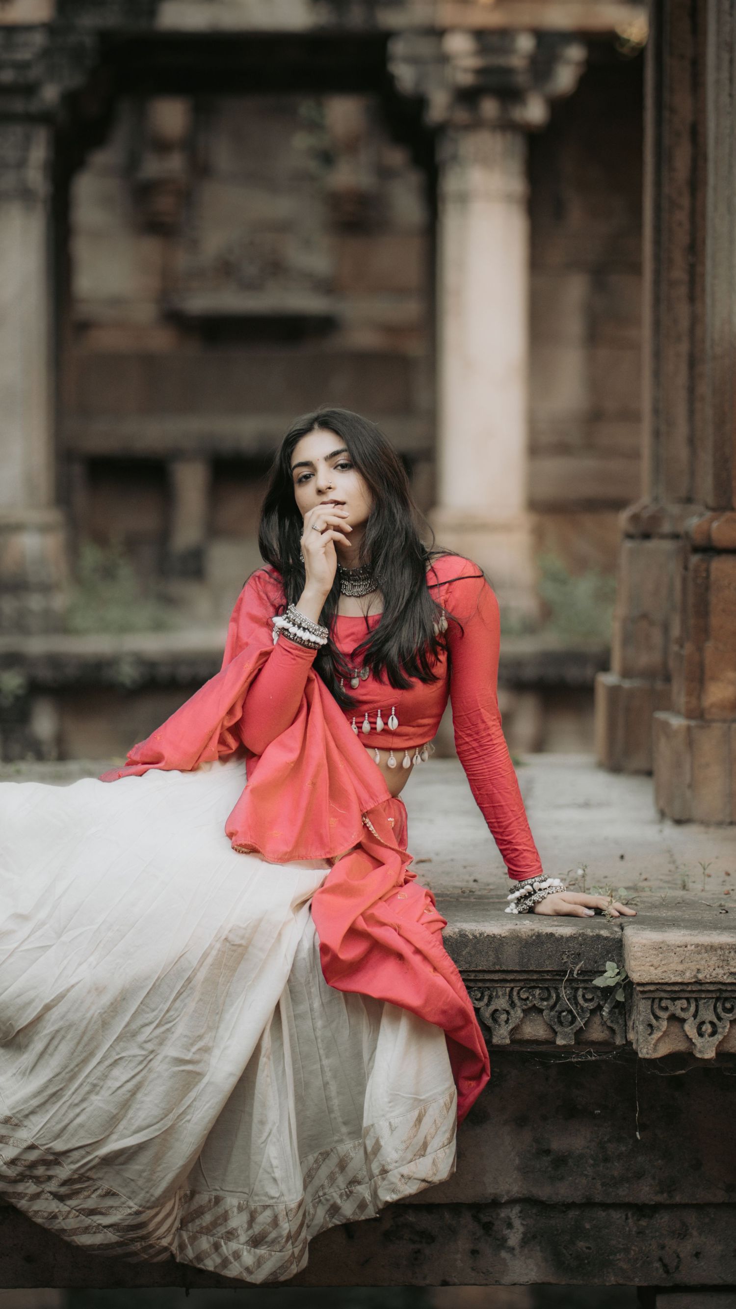Young woman in red and white traditional Indian attire sits pensively on ancient temple steps.