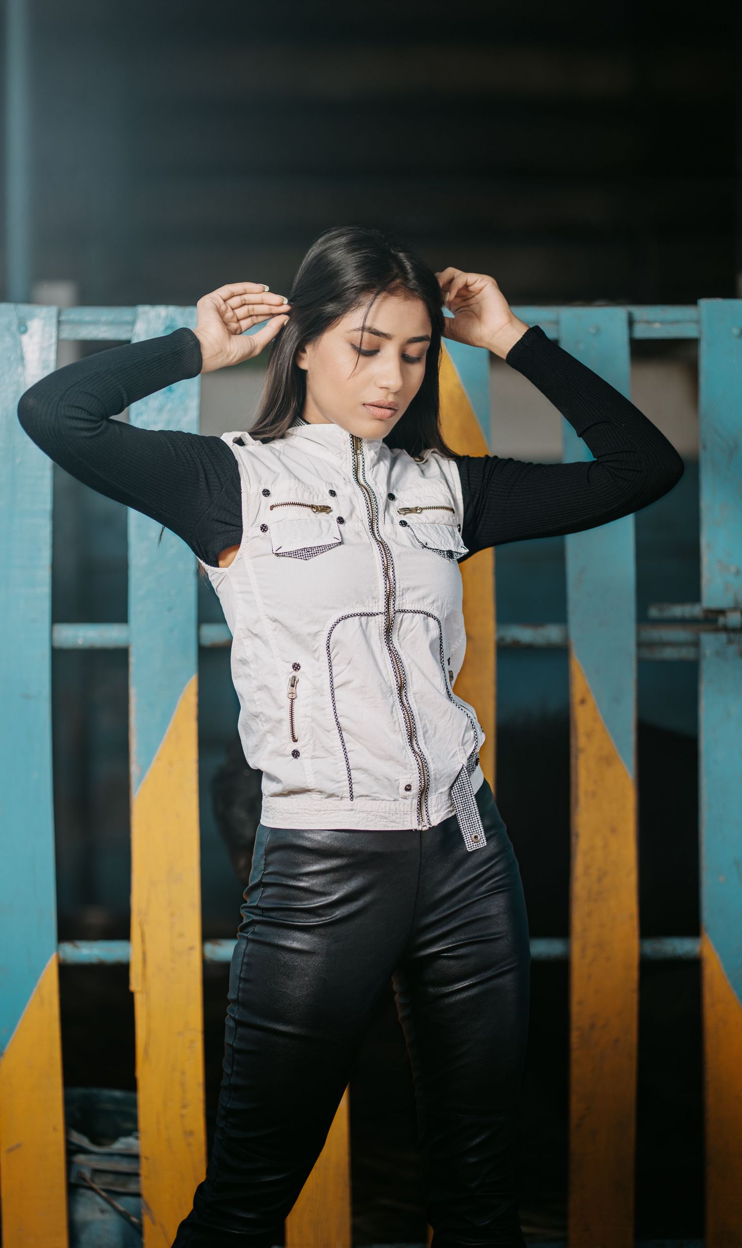Young woman posing in white vest jacket, black turtleneck, and leather pants against colorful wooden background.