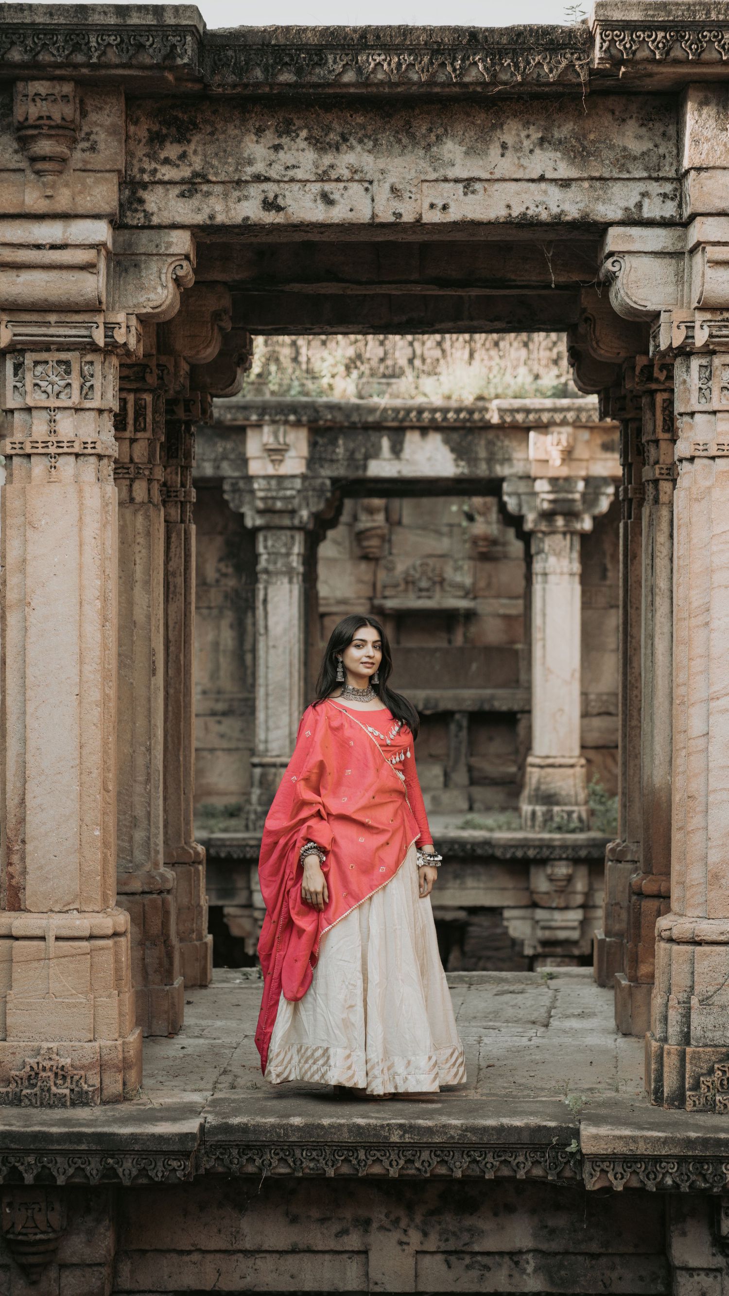 Woman in white lehenga and red dupatta posing at ancient stone temple archway in India.