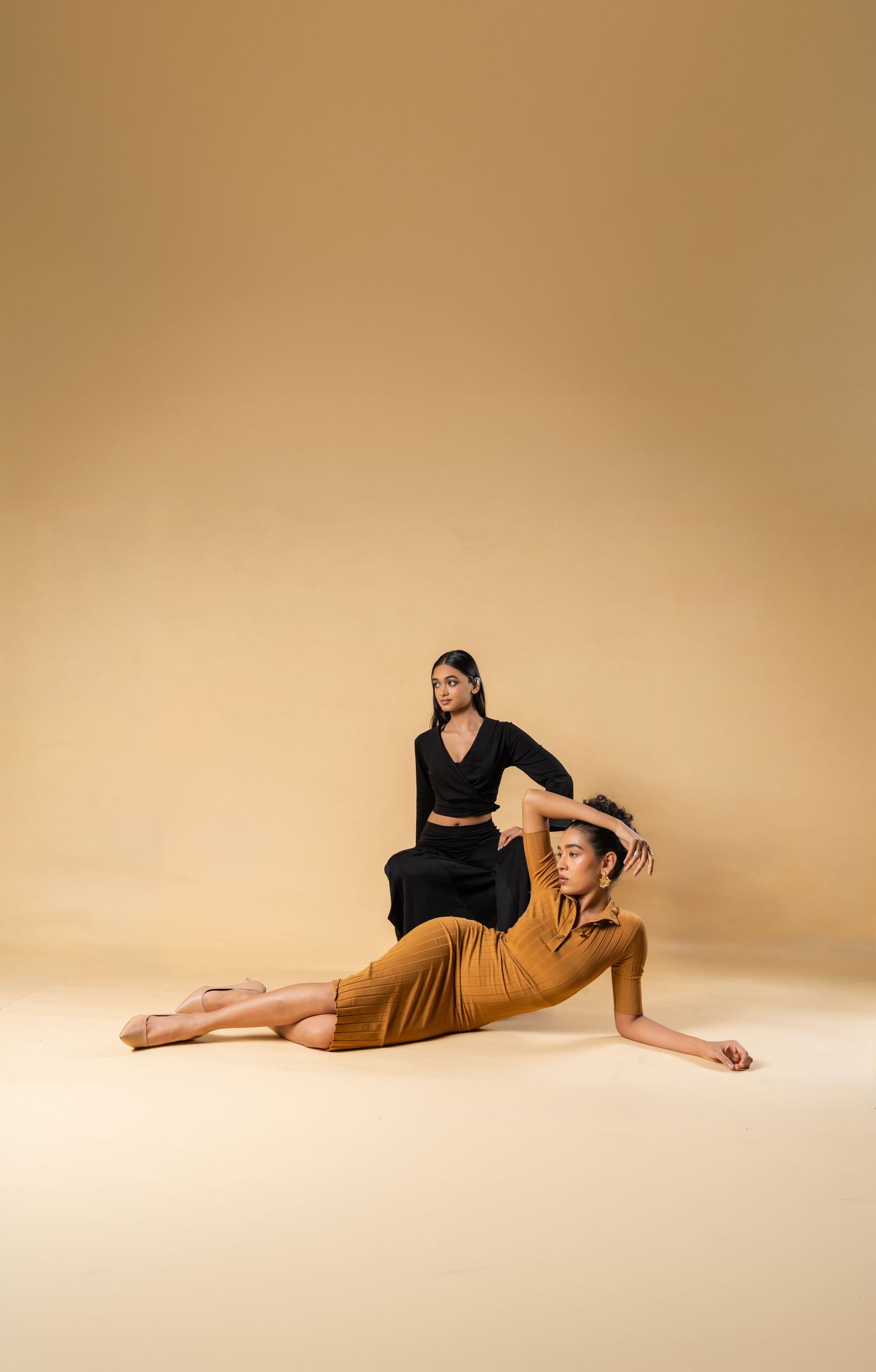 Two women pose gracefully in a studio, one in black wrap top and skirt, other in mustard dress on the floor.
