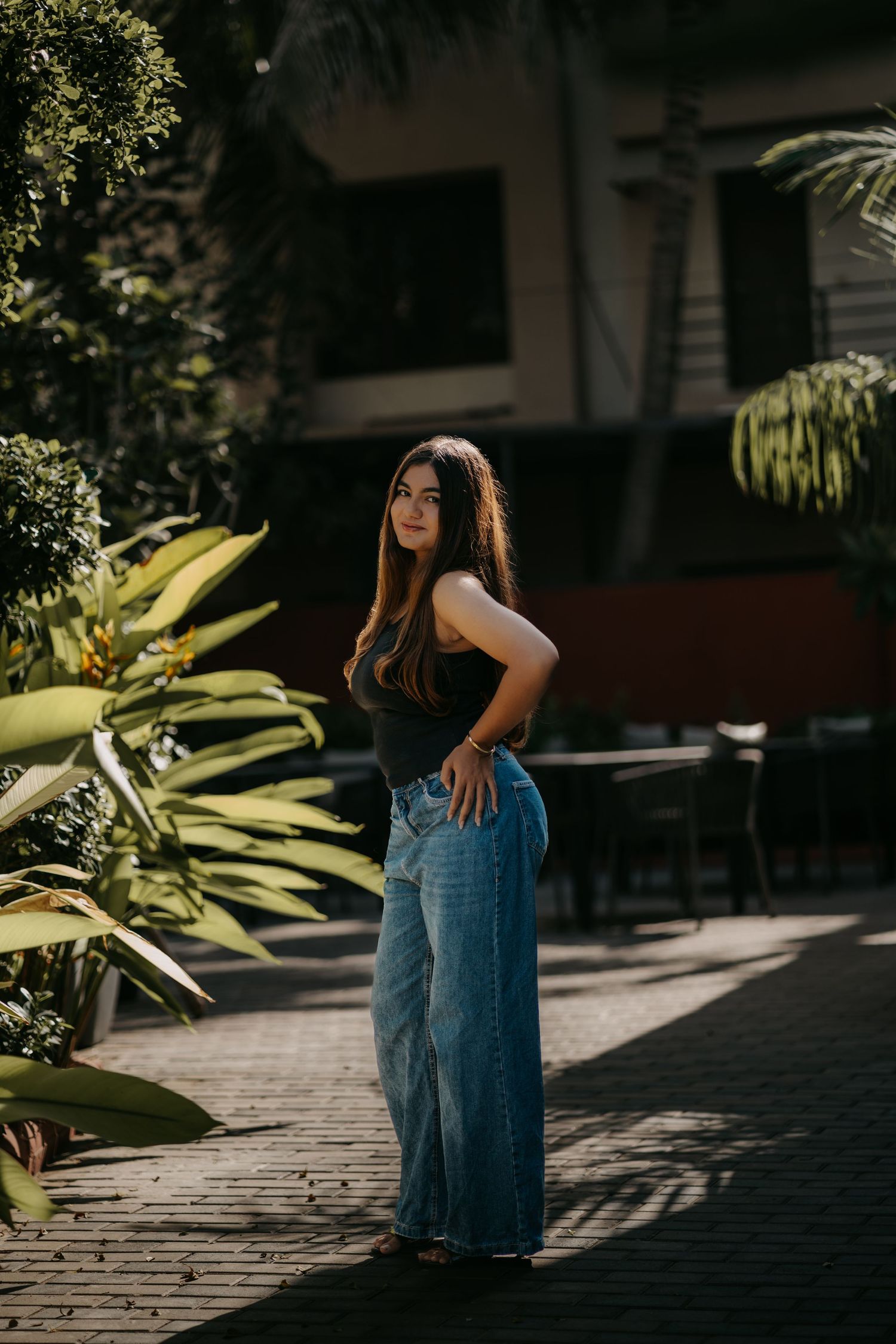 Young woman in wide-leg jeans and black top posing outdoors among tropical plants on a sunny day.