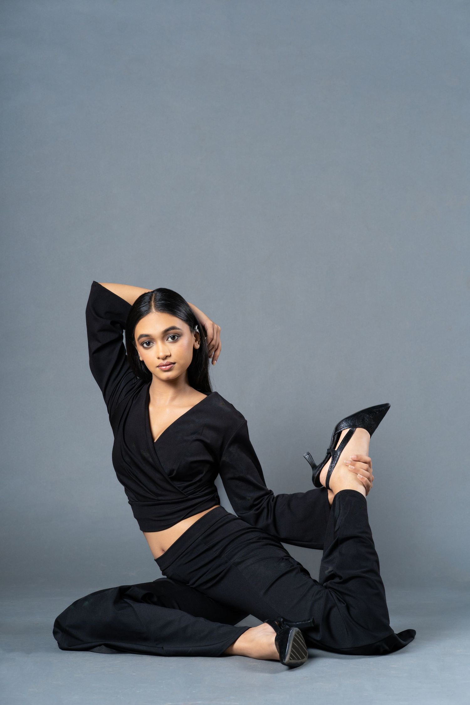 A young woman in a black wrap top and pants poses gracefully on the floor with one leg raised, against a gray background.