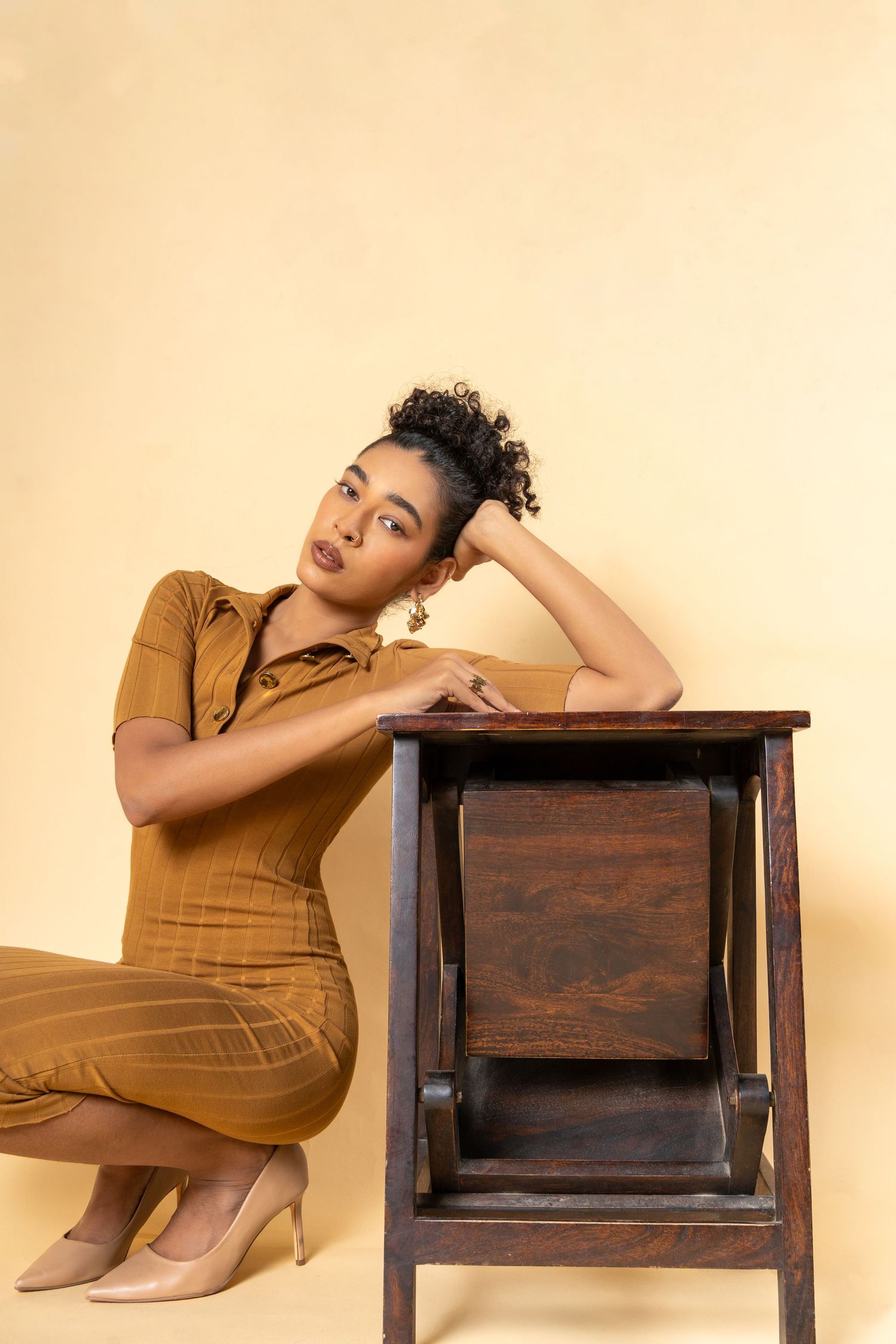 A stylish woman in a brown jumpsuit poses gracefully beside a wooden cabinet against a beige background.