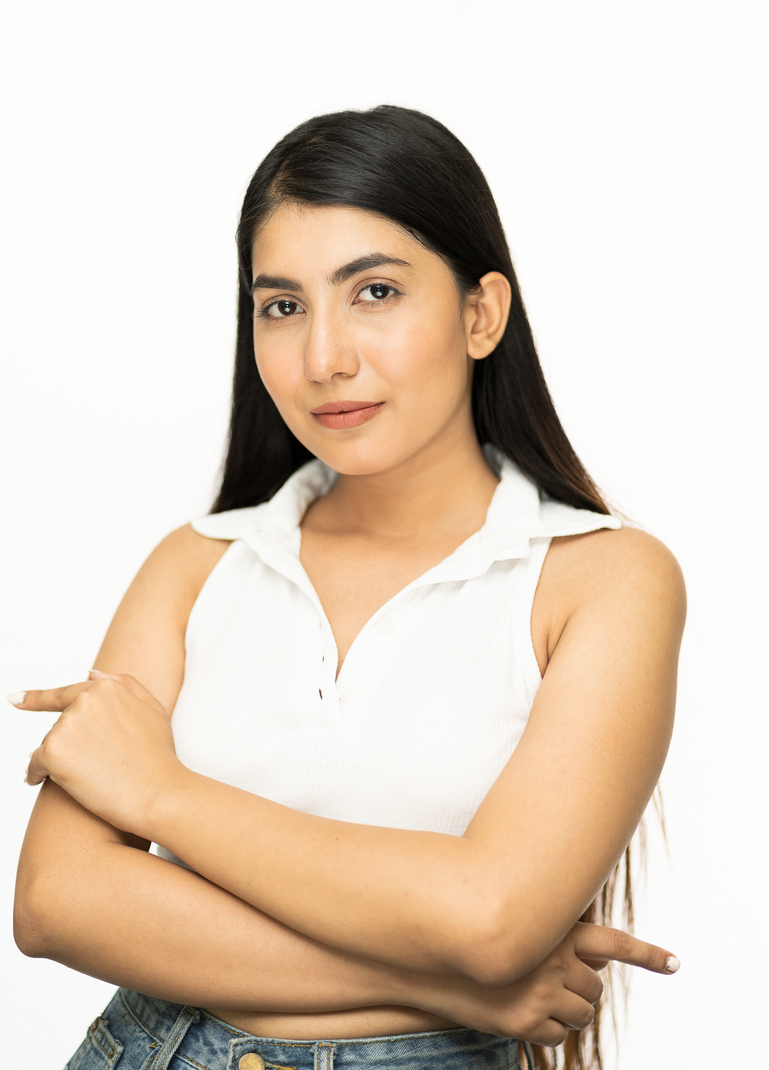 Young woman with long dark hair, arms crossed, wearing a white sleeveless polo top and jeans, posing against white background.